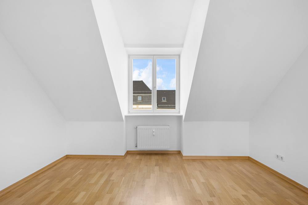 Leeres Zimmer mit Dachschräge, Holzfußboden und Fenster mit Blick auf Häuser.