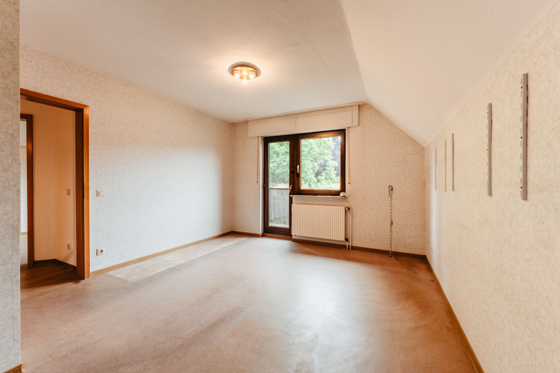 Leeres Zimmer mit Fenster, Tür, Heizung und heller Deckenleuchte, Blick auf grüne Bäume draußen.