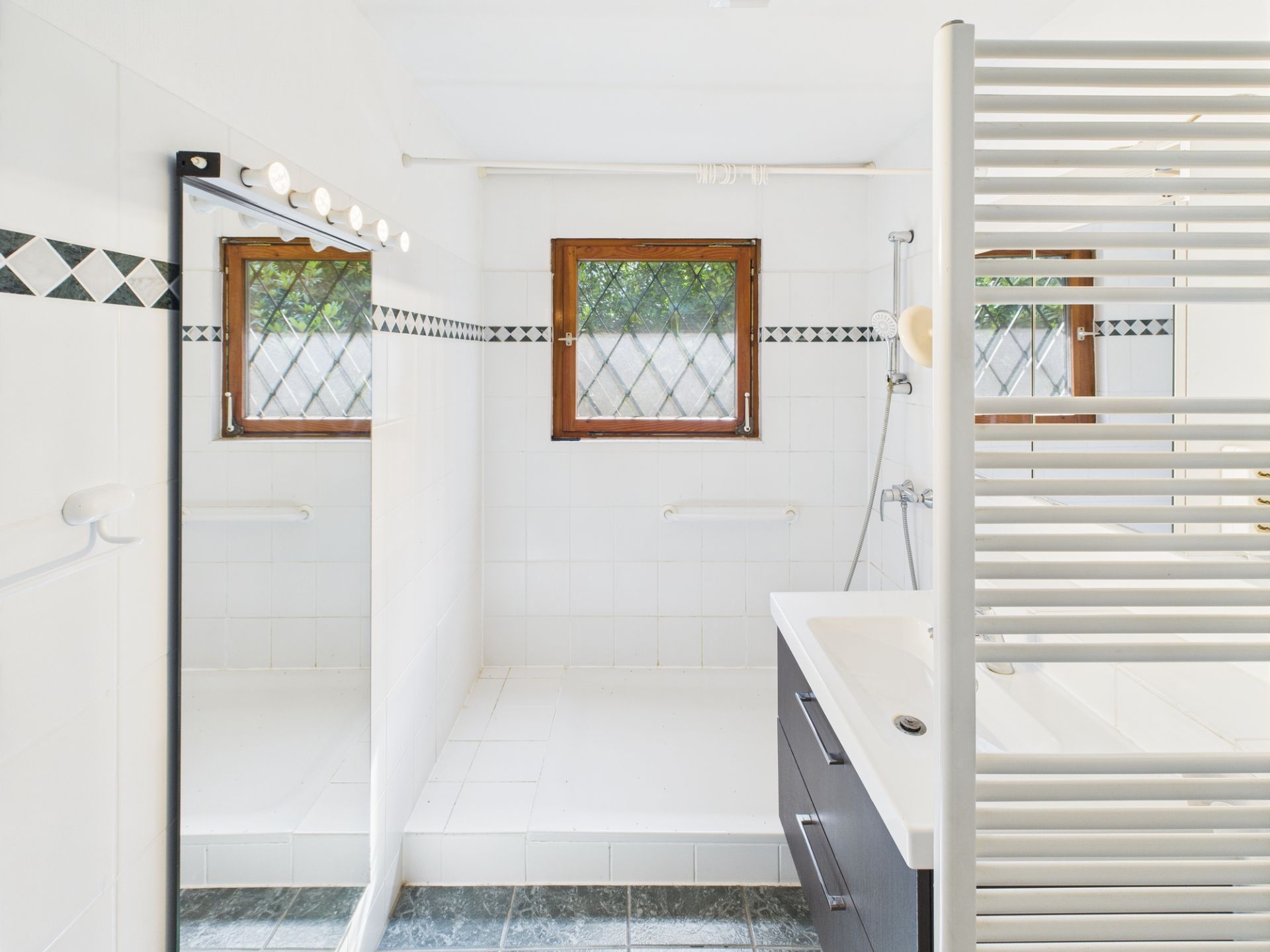 Modernes, weiß gefliestes Badezimmer mit Dusche, zwei Fenstern und großem Spiegel.