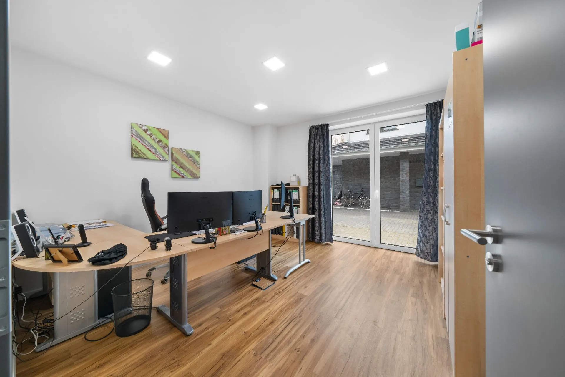 Helles Arbeitszimmer mit zwei Schreibtischen, Monitoren, Regal und Fenstertür zum Innenhof.