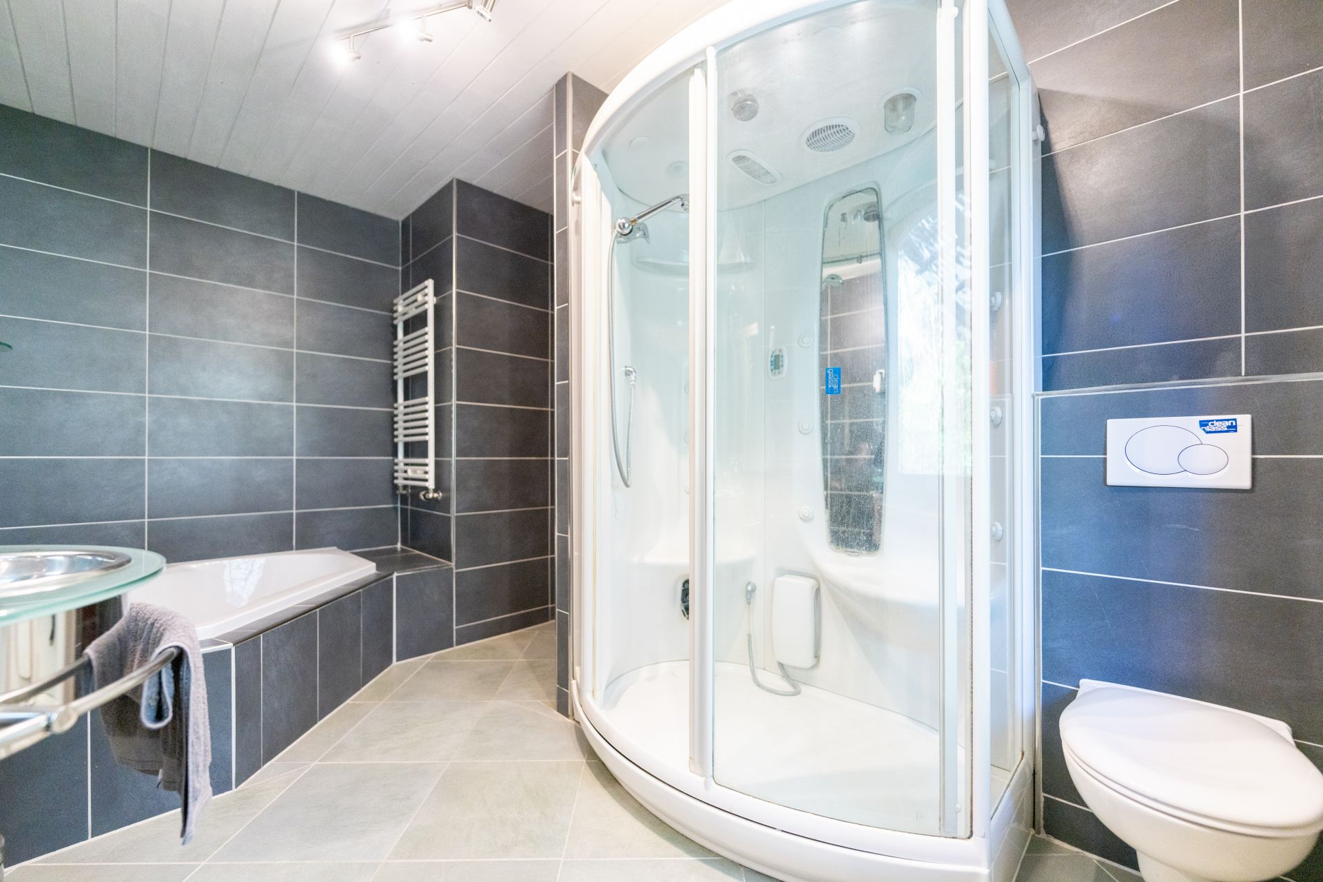 Modernes Bad mit Dusche, Badewanne, Wandtoilette und Handtuchheizkörper, alles in grauen Fliesen gehalten.