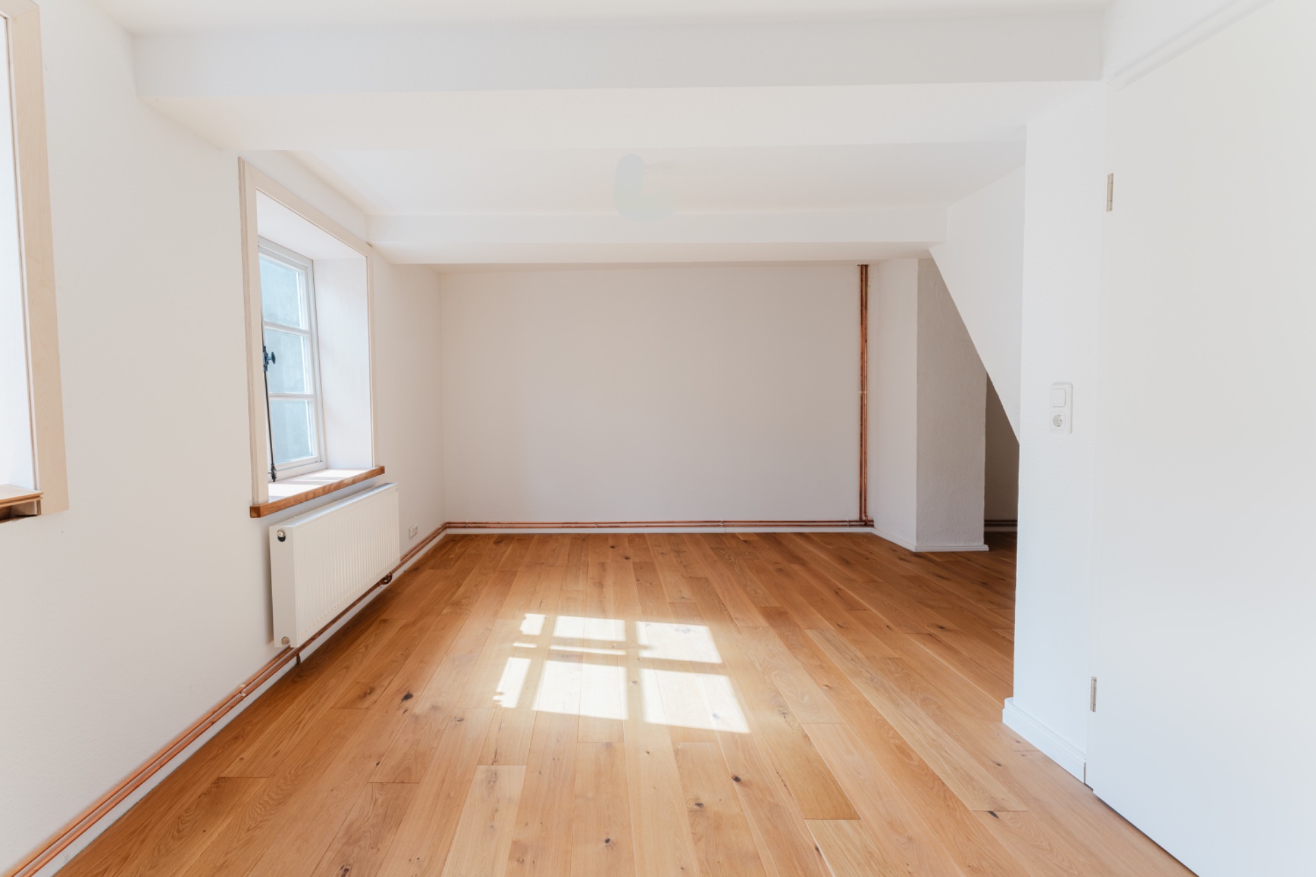 Heller leerer Raum mit Holzfußboden, weiß gestrichenen Wänden und Fenster auf der linken Seite.