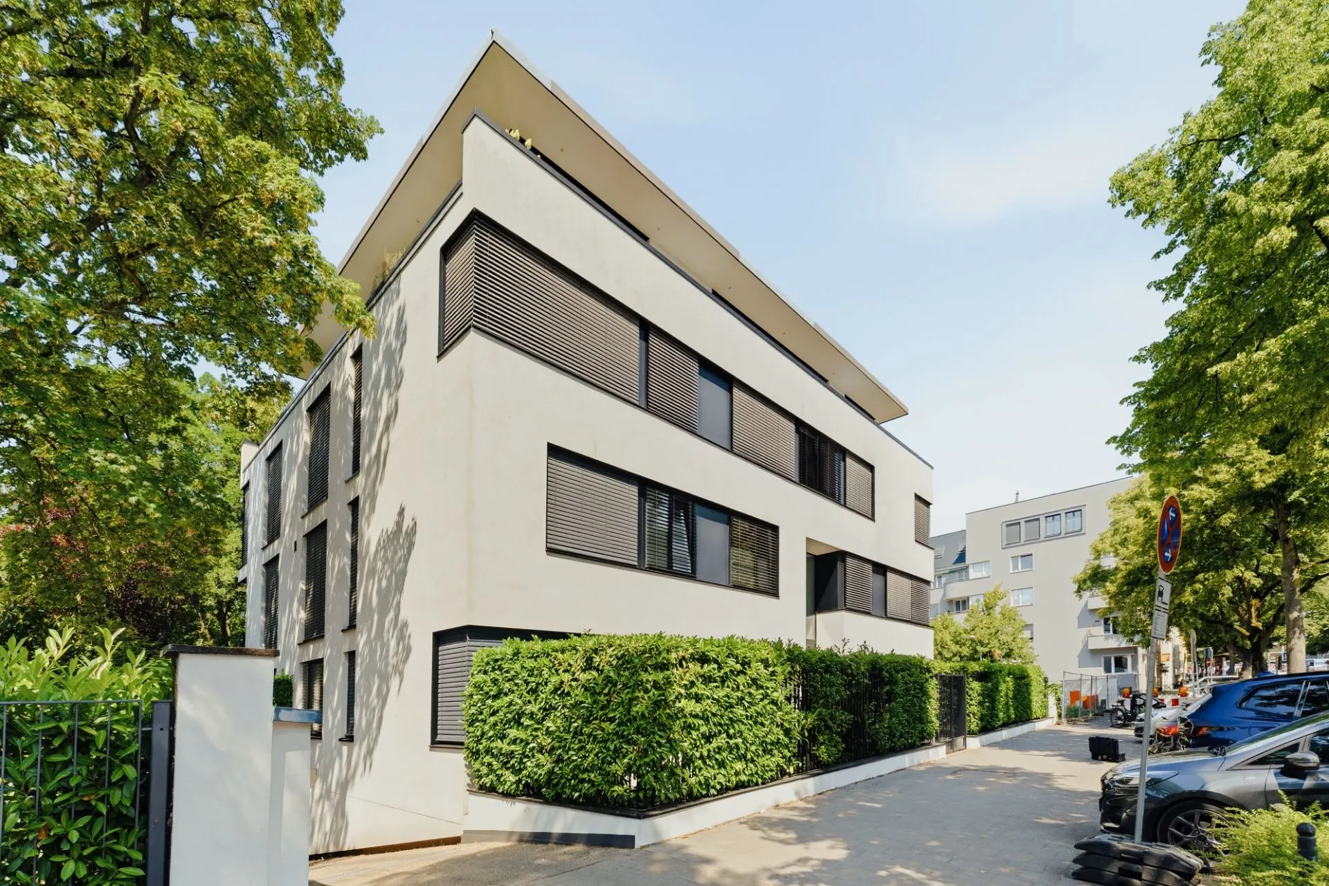 Modernes Wohngebäude mit großen Fenstern und Rollläden, umgeben von Bäumen und Hecken.
