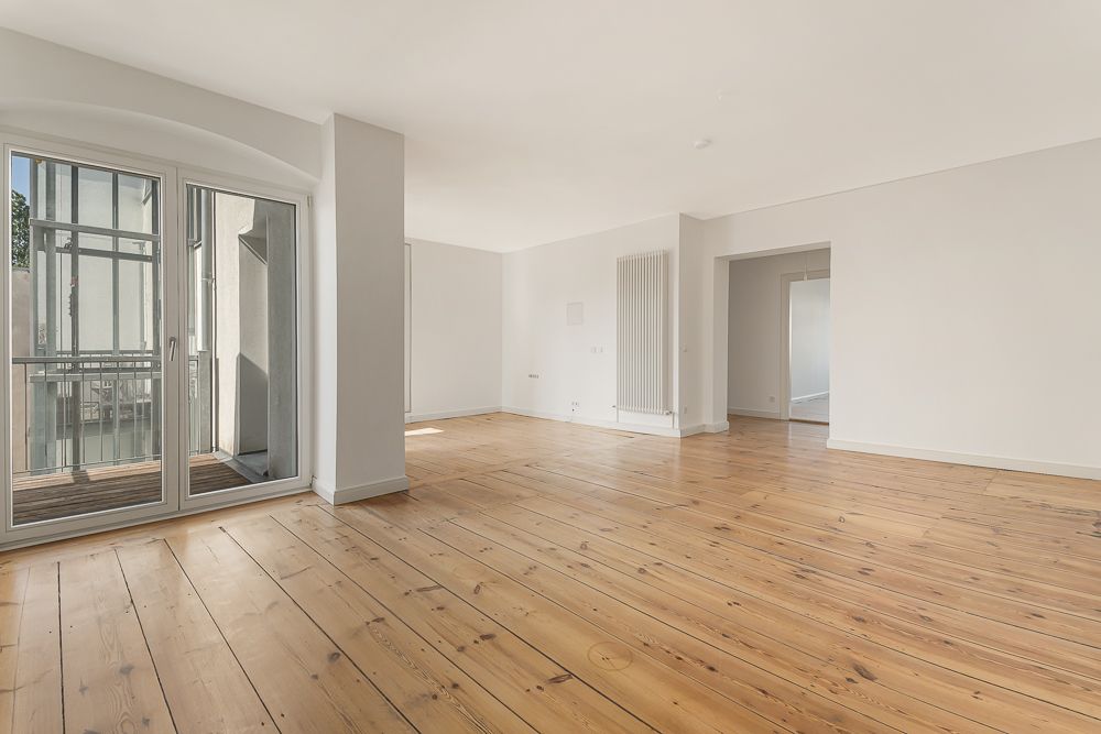 Helles, leeres Zimmer mit Holzboden, weißen Wänden und großer Glastür zum Balkon.