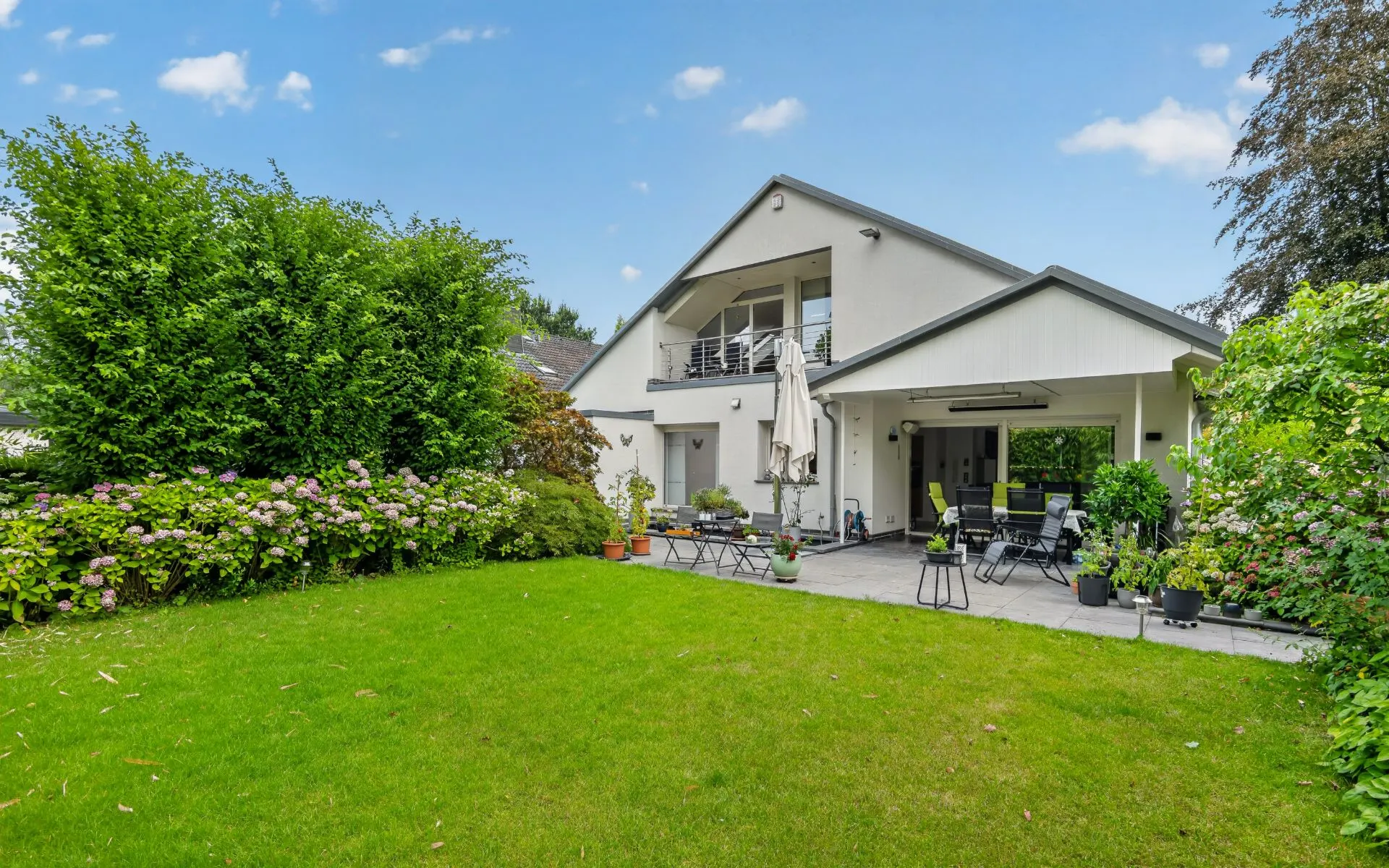 Moderner Garten mit Terrasse, Stühlen, Tisch und vielen Pflanzen vor einem Einfamilienhaus.