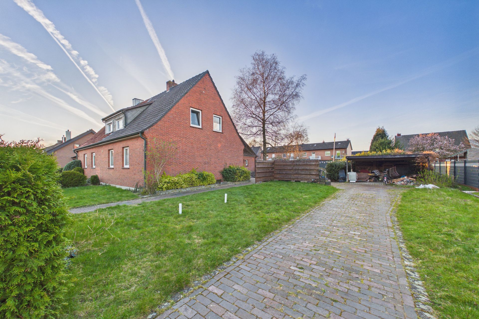 Backsteinwohnhaus mit schrägem Dach, gepflasterte Einfahrt und Garten im Vordergrund.