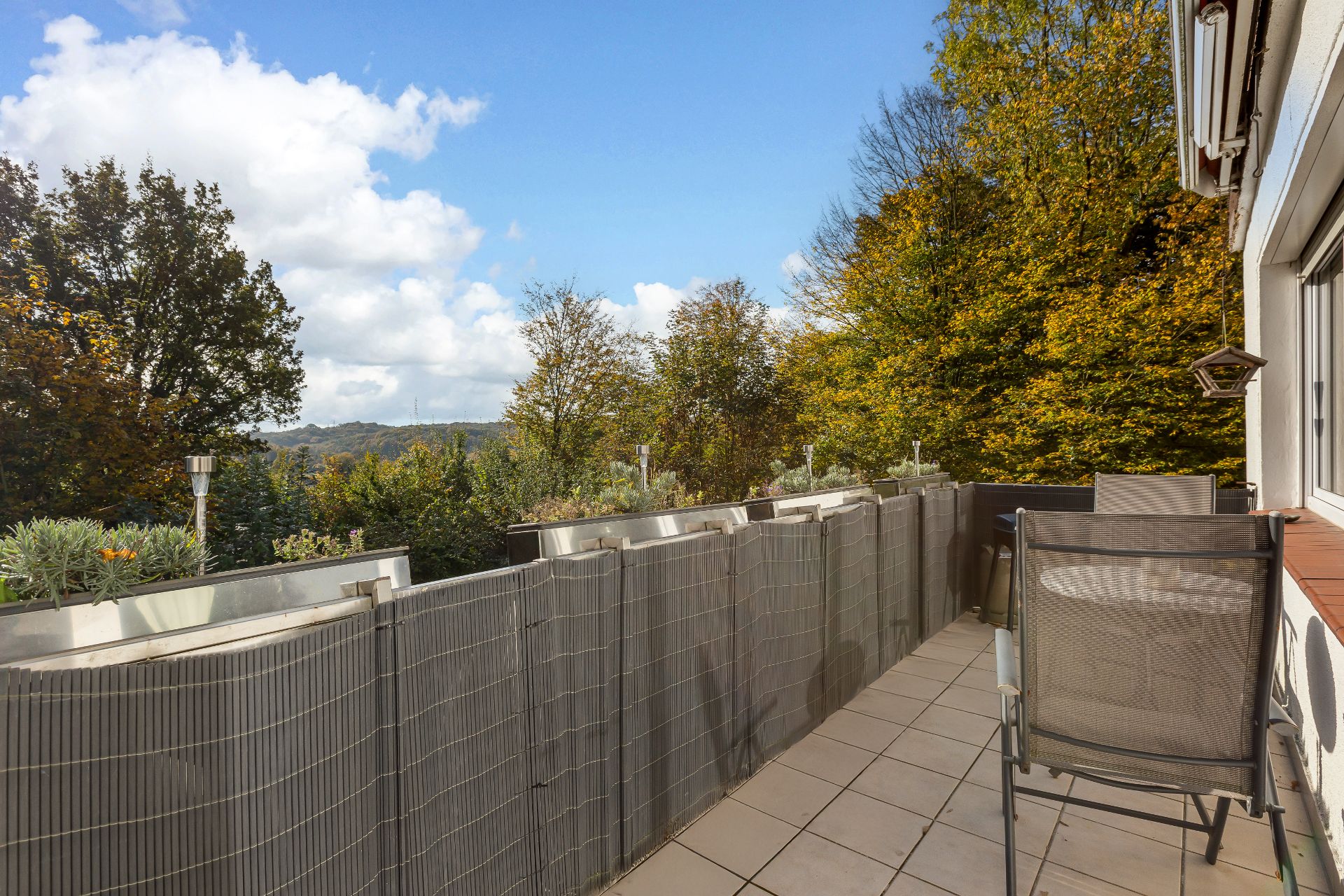 Balkon mit zwei Stühlen, Tisch und Blick auf Bäume und Hügel unter blauem Himmel.