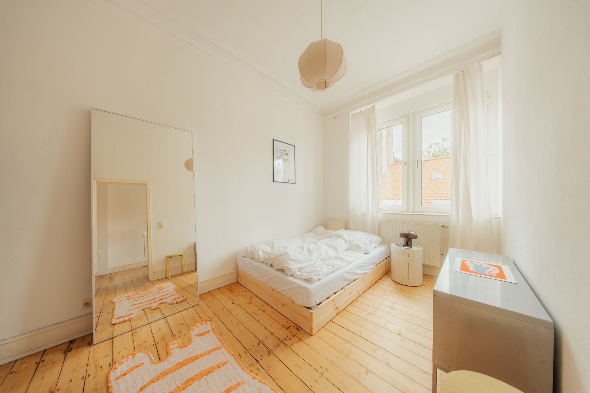 Helles Schlafzimmer mit Holzboden, Bett, Spiegel und Fenster mit weißen Vorhängen.