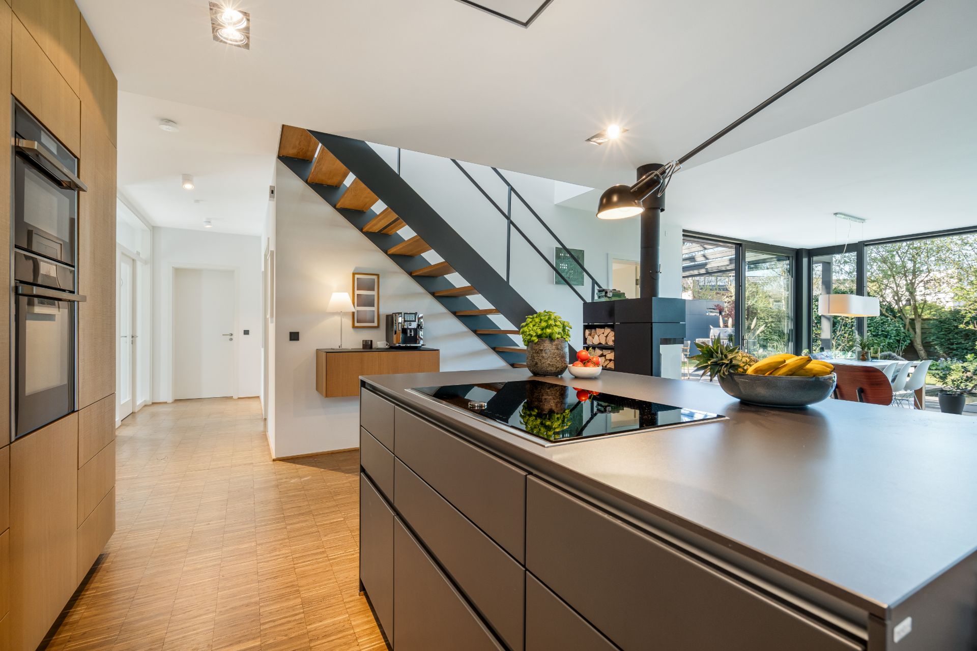 Moderne offene Küche mit Kochinsel, Treppe und Blick auf Essbereich mit großen Fenstern.