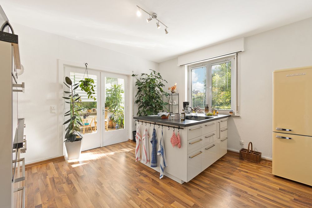 Helle Küche mit Kücheninsel, gelbem Kühlschrank, Pflanzen und großen Fenstern.