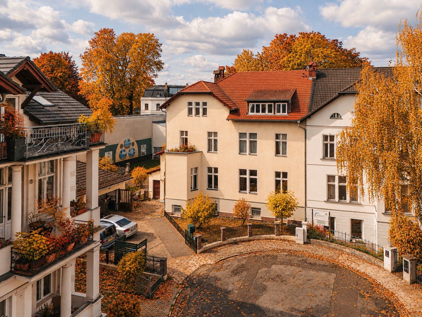 Helles Wohnhaus mit rotem Dach, Bäumen mit Herbstlaub und kleinem Hof mit geparktem Auto.