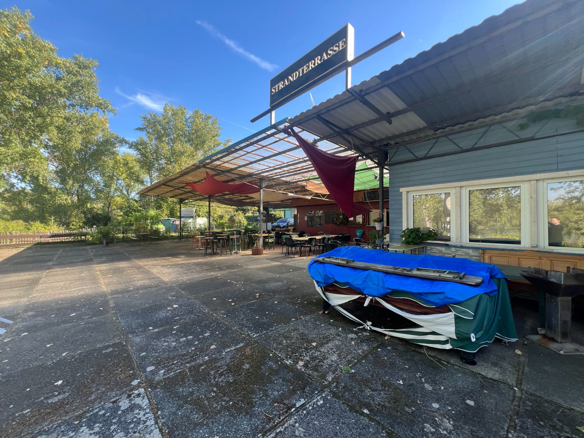 Überdachte Terrasse mit Tischen, Stühlen und einem Boot, daneben Fensterfront und Bäume im Hintergrund.