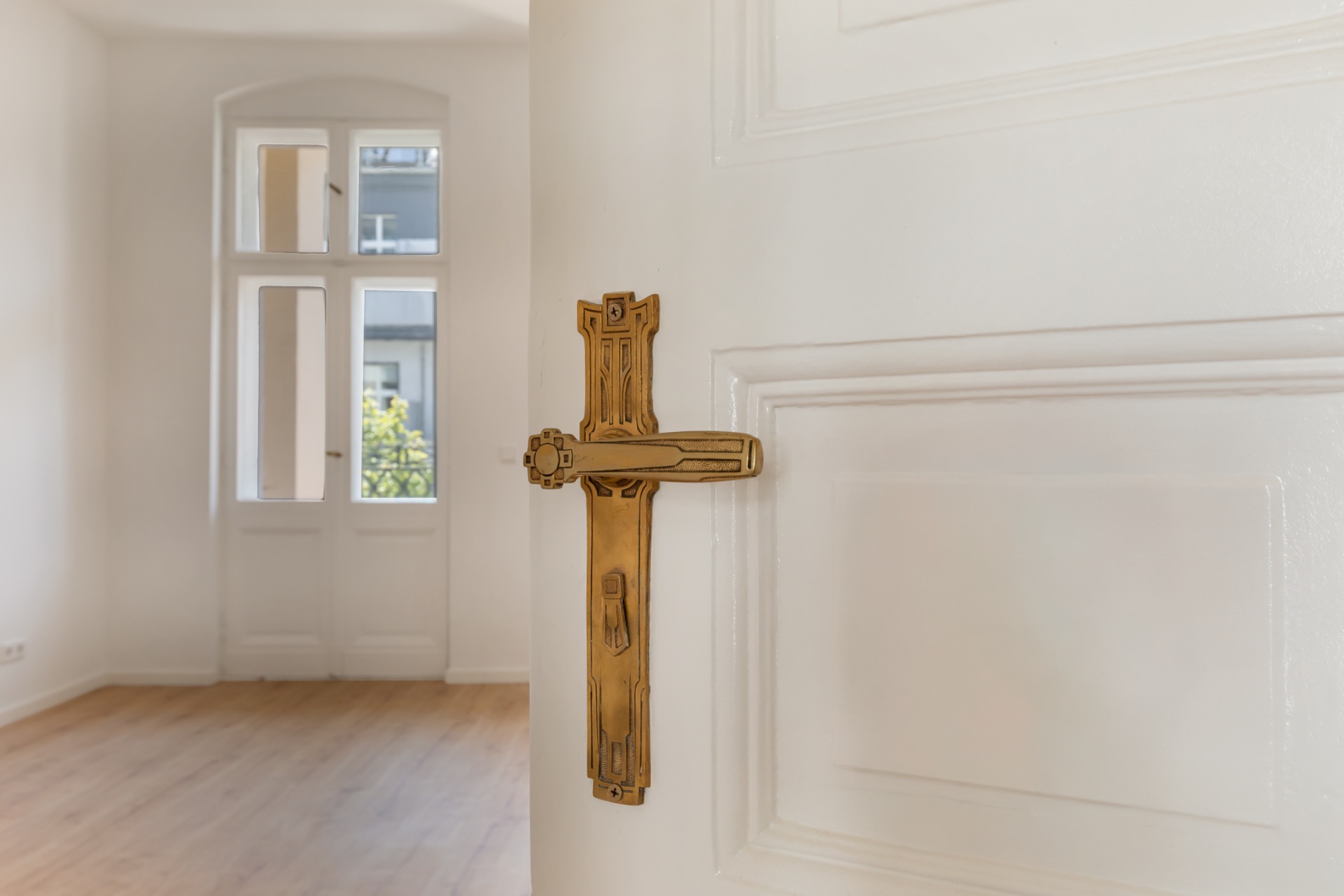 Weiße Tür mit goldenem Griff, dahinter ein heller Raum mit Holzboden und Fenstern.
