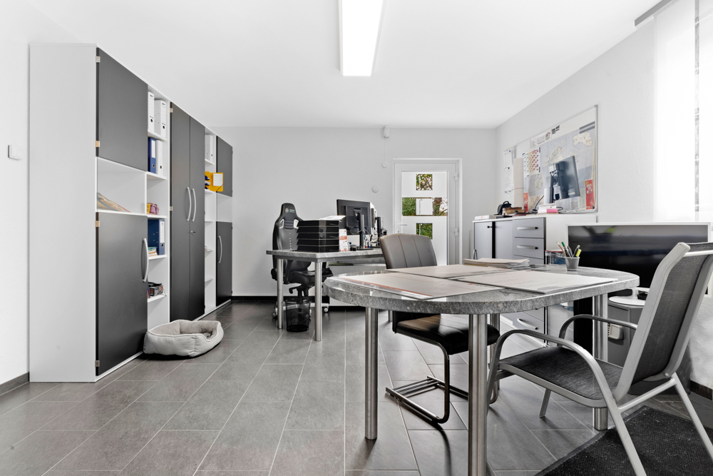 Modernes Arbeitszimmer mit Schreibtisch, Stühlen, Regalen und Hundebett auf grauem Fliesenboden.