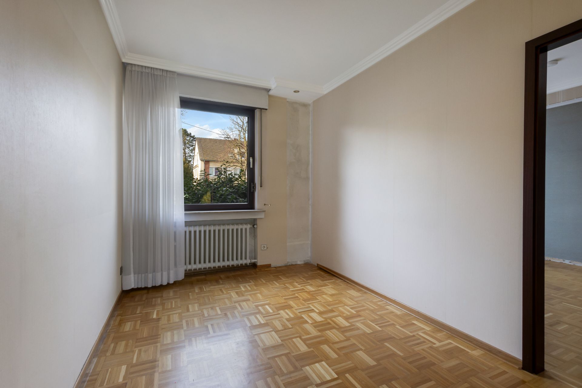 Leeres Zimmer mit Parkettboden, Fenster mit weißen Vorhängen und Blick auf Nachbarhäuser.