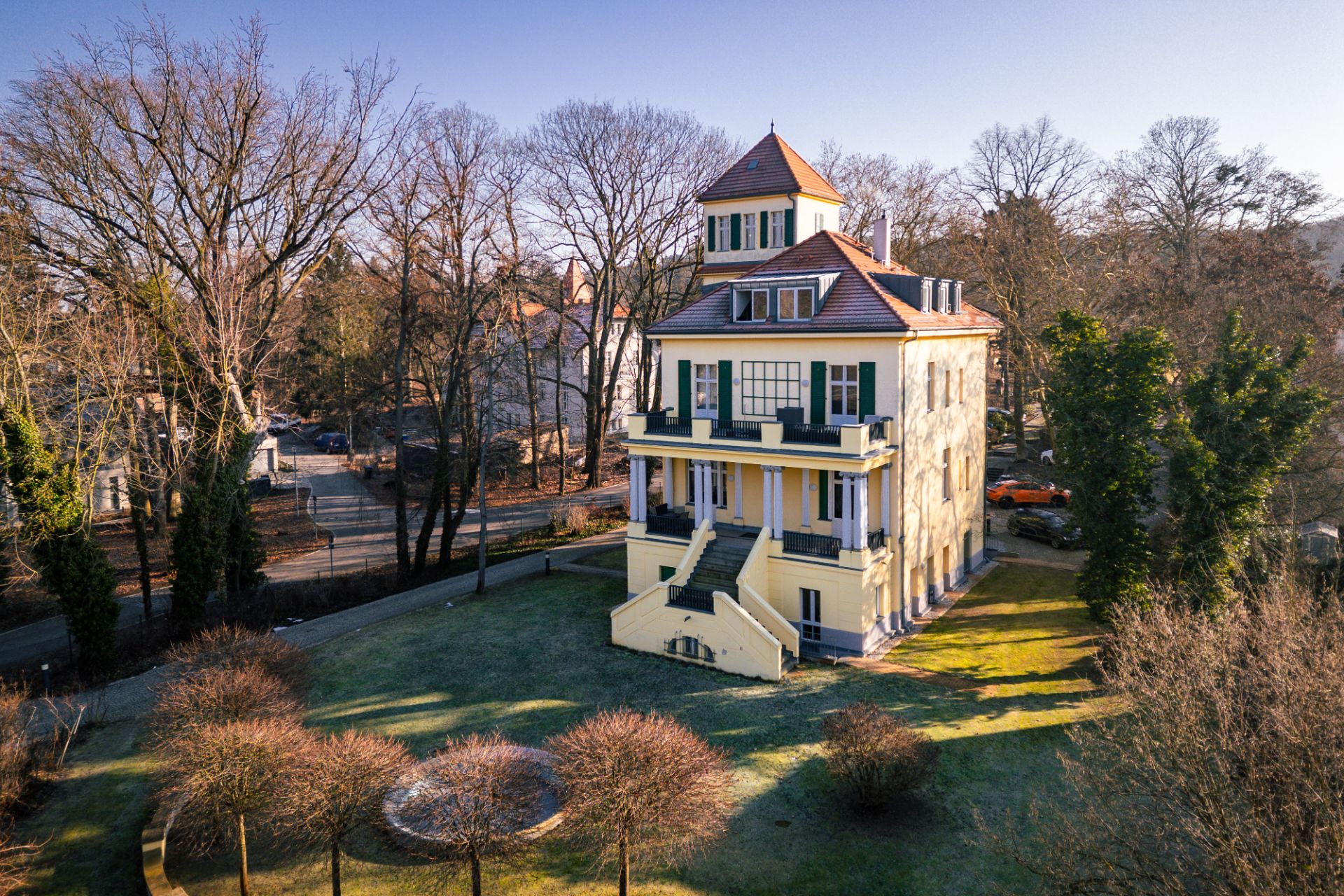 Großes, gelbes Wohnhaus mit mehreren Etagen und Balkon, umgeben von Bäumen und Garten.