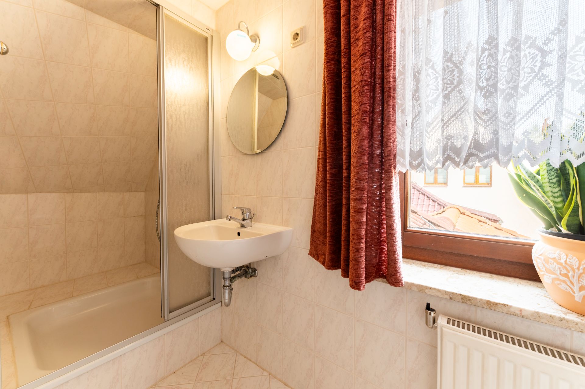Kleines Badezimmer mit Dusche, Waschbecken, rundem Spiegel, Fenster und Topfpflanze.