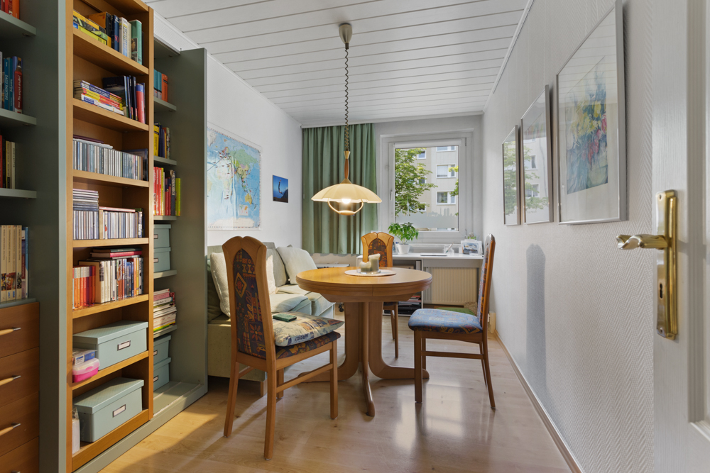 Heller Essbereich mit rundem Tisch, vier Stühlen, Bücherregal und großem Fenster.