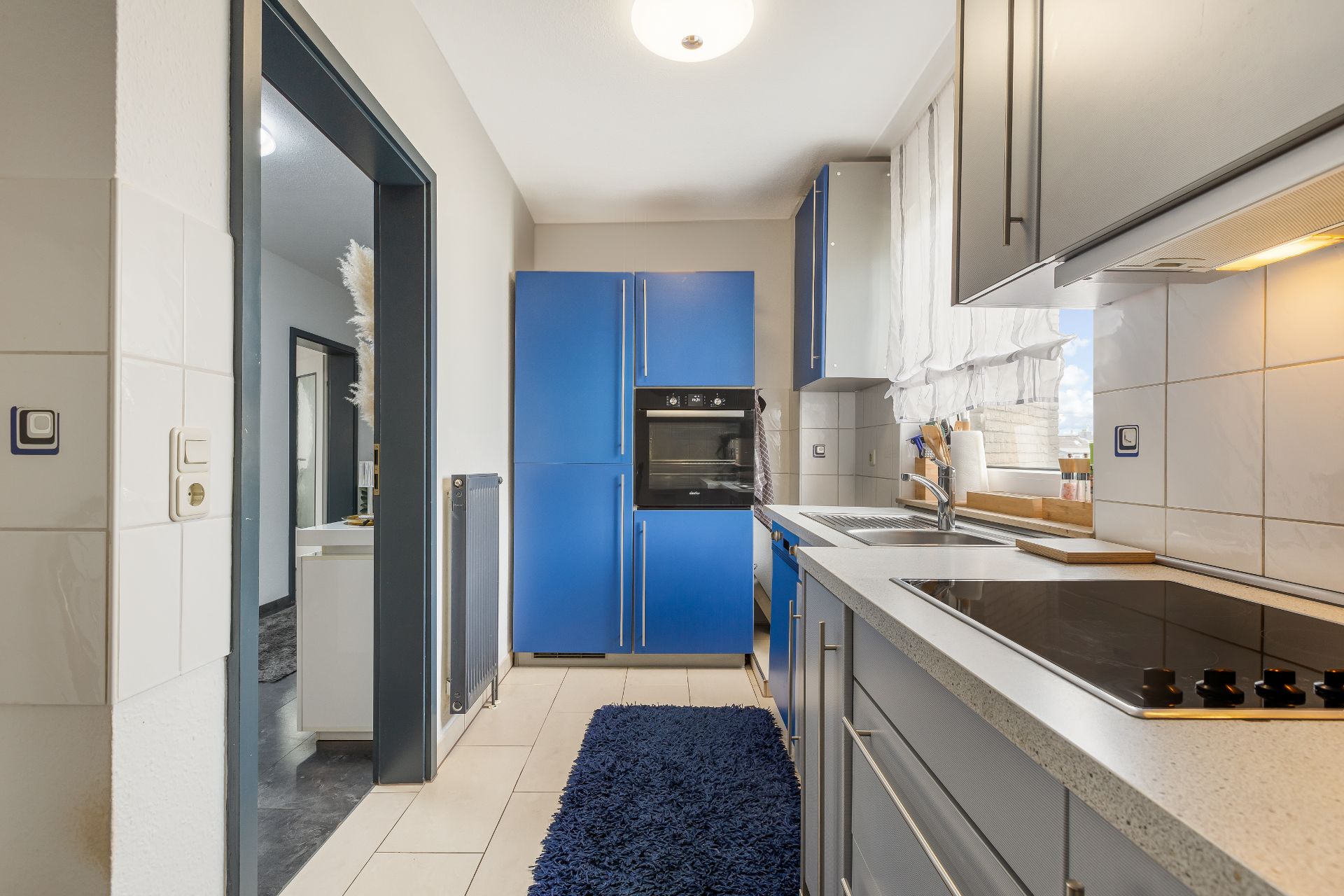 Moderne Küche mit blauen Schränken, Herd, Spüle und großem Fenster mit Gardine.