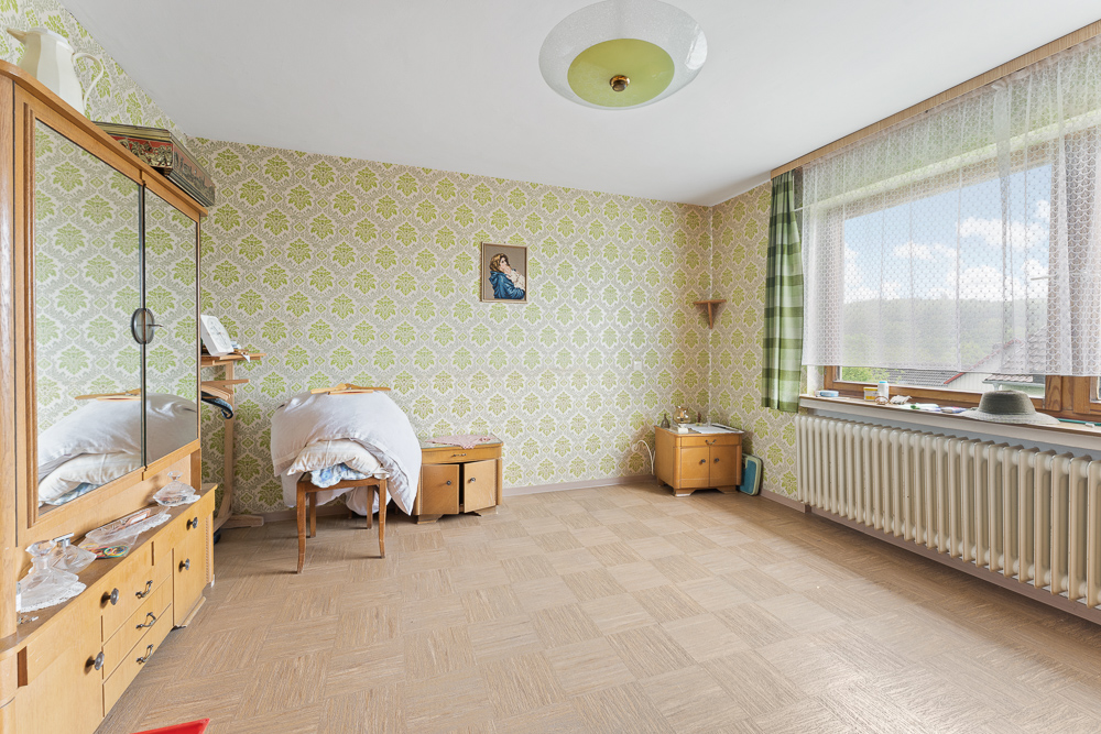 Helles Zimmer mit gemusterter Tapete, Holzschrank, Stuhl, Nachttisch und großem Fenster mit Gardinen.