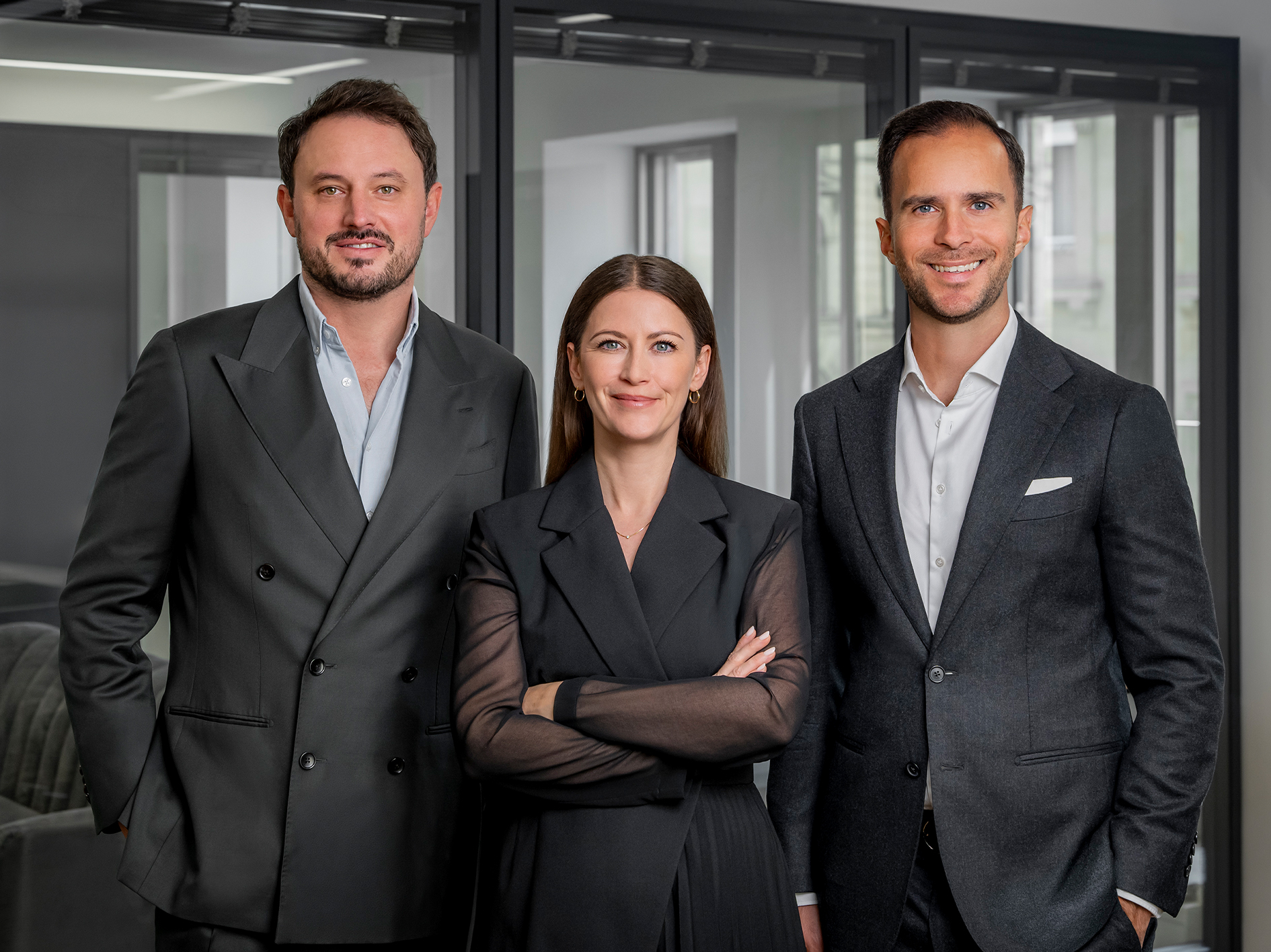 Drei Personen in eleganter Kleidung stehen in einem modernen Büroraum mit Glaswänden.