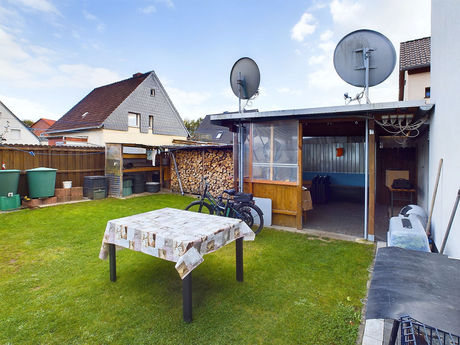 Garten mit Tisch, Fahrrädern, Holzstapel und Schuppen mit Satellitenschüsseln.