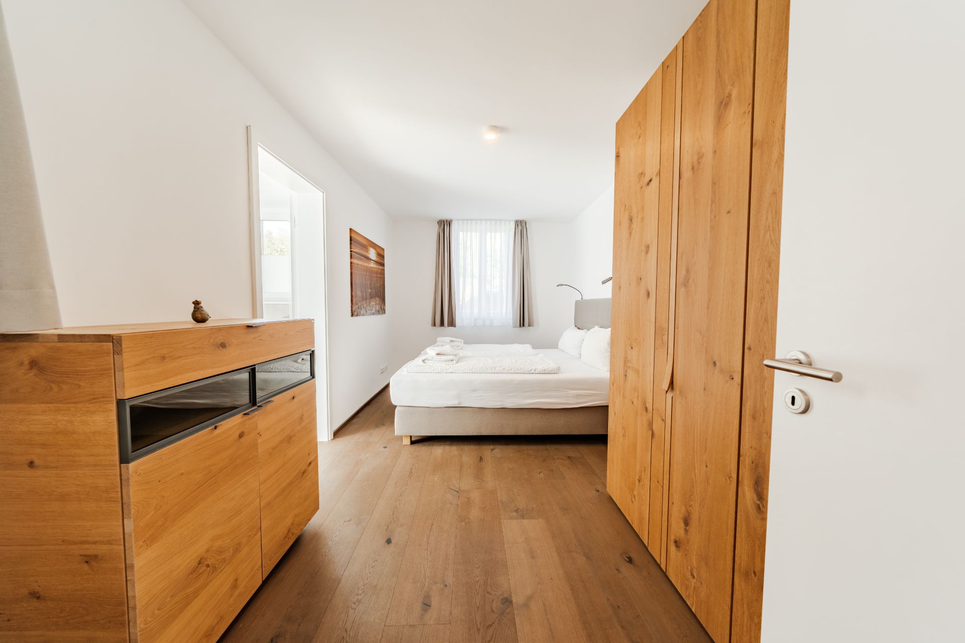 Helles Schlafzimmer mit Holzschrank, Kommode und Doppelbett, Fenster mit Vorhängen im Hintergrund.