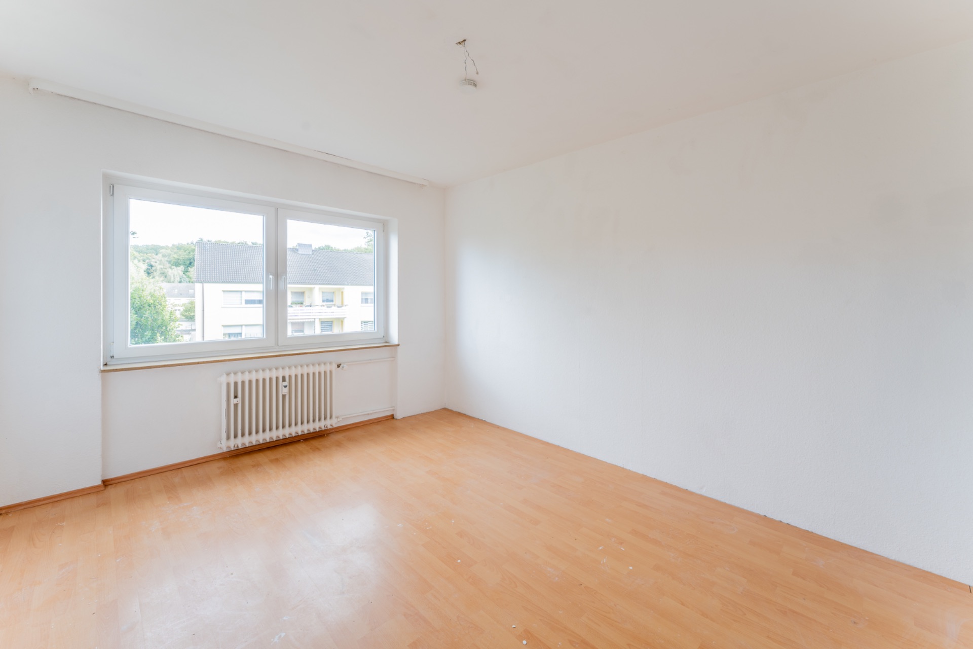 Leerer, heller Raum mit weißer Wand, großem Fenster und Holzfußboden.