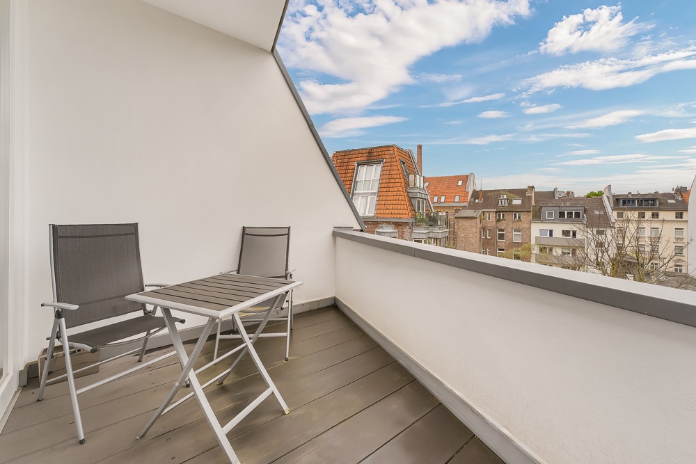 Balkon mit zwei Stühlen und einem Tisch, Blick auf Nachbarhäuser und blauen Himmel.