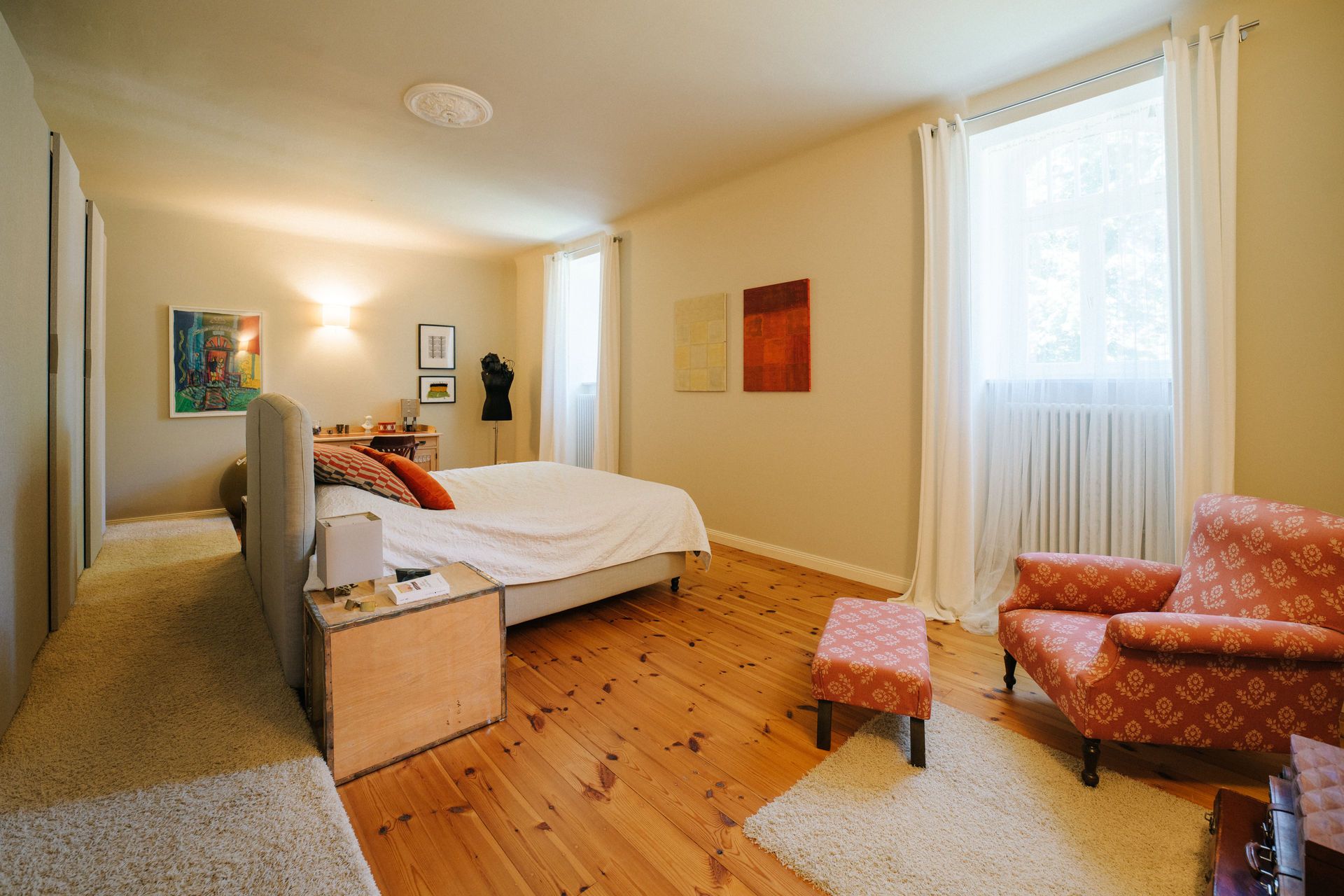 Helles Schlafzimmer mit Doppelbett, zwei Fenstern, Teppichen und gemütlichem Sessel mit Hocker.