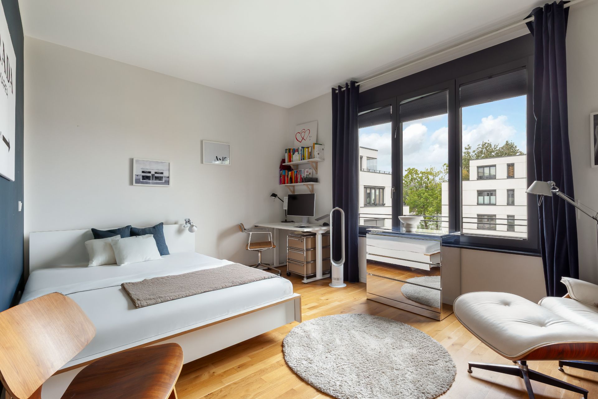 Helles Schlafzimmer mit Schreibtisch, großem Fenster, Bett und Sessel auf Holzboden.