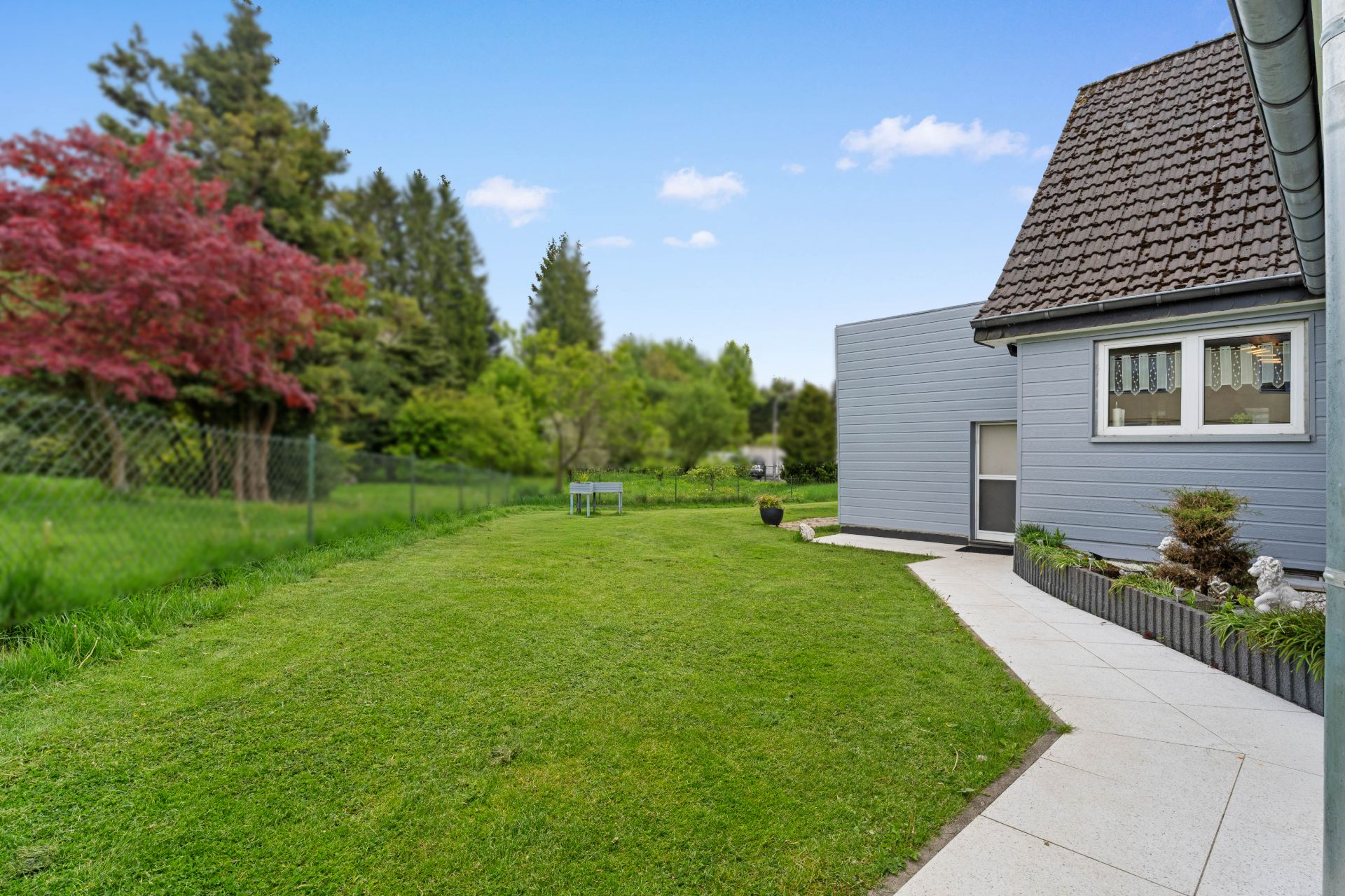 Grüner Garten mit Rasen, Bäumen und Hausfassade rechts, Weg entlang des Hauses.