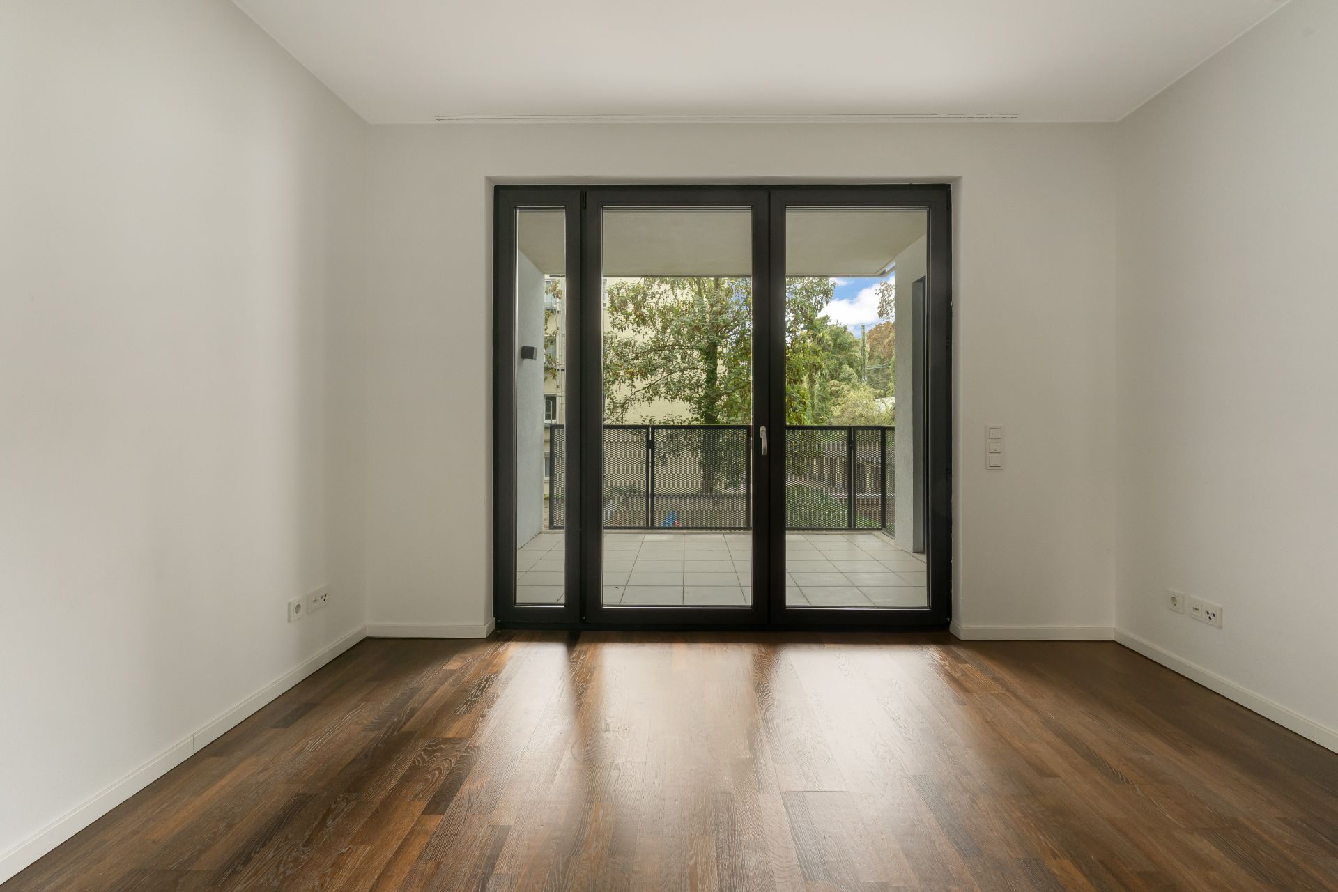 Leerer Raum mit Holzboden und großer Glastür zu einem Balkon mit Blick auf Bäume.