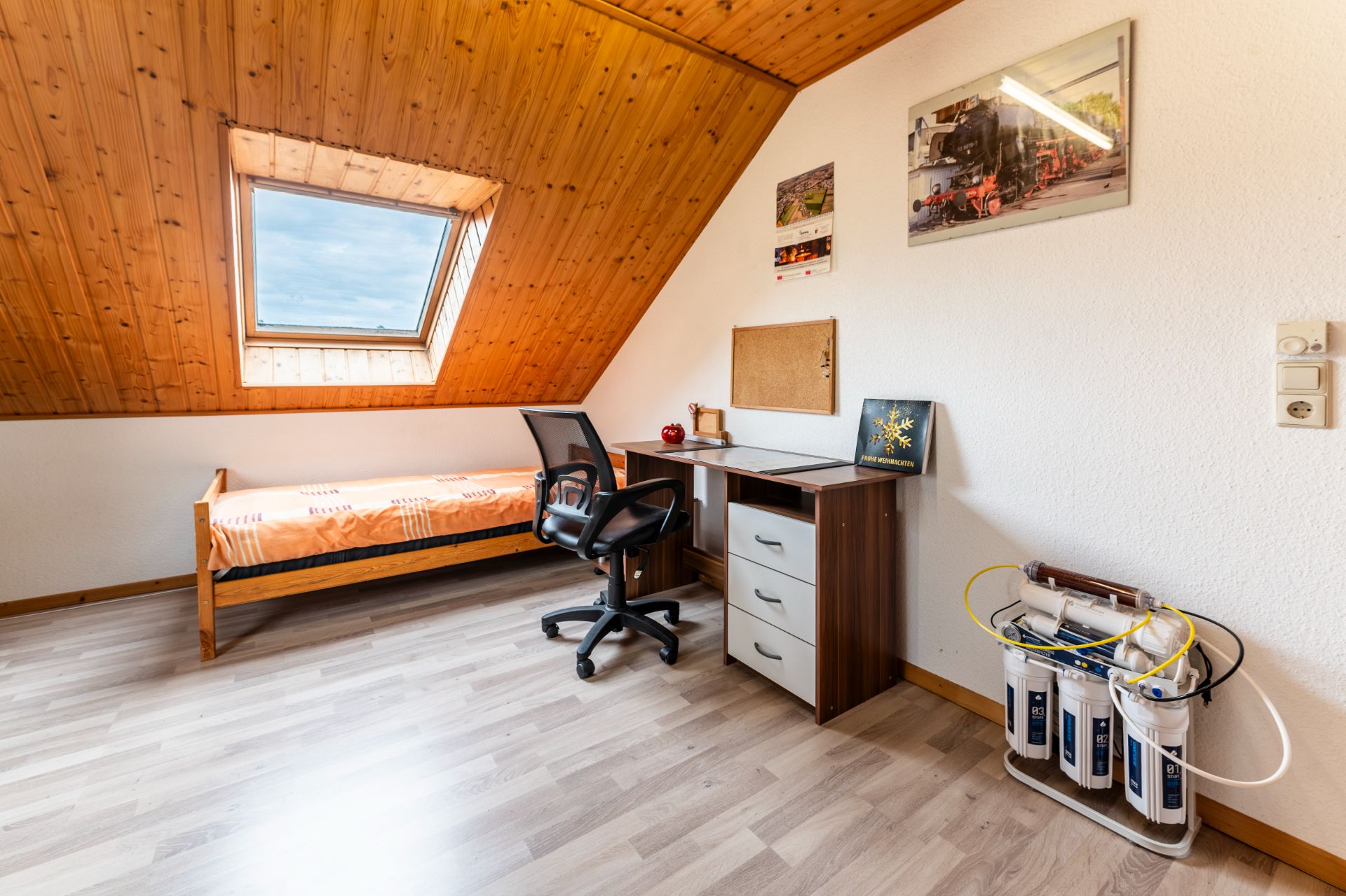 Dachzimmer mit Bett, Schreibtisch, Stuhl und Wasserfilter. Fenster in der Schräge.