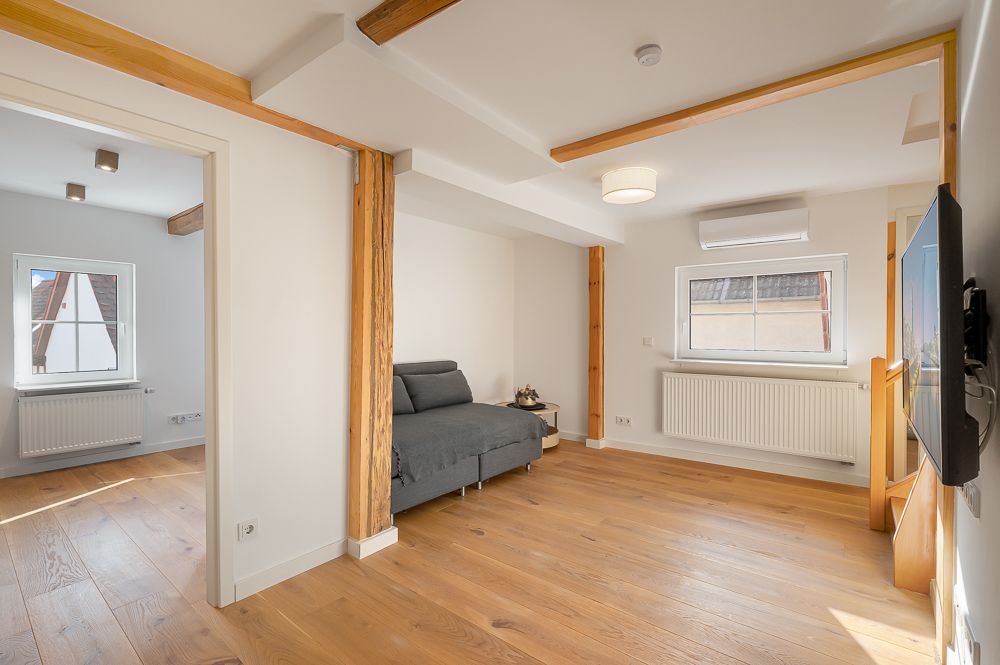 Helles Wohnzimmer mit Holzboden, Schlafsofa, TV an der Wand und sichtbaren Holzbalken.