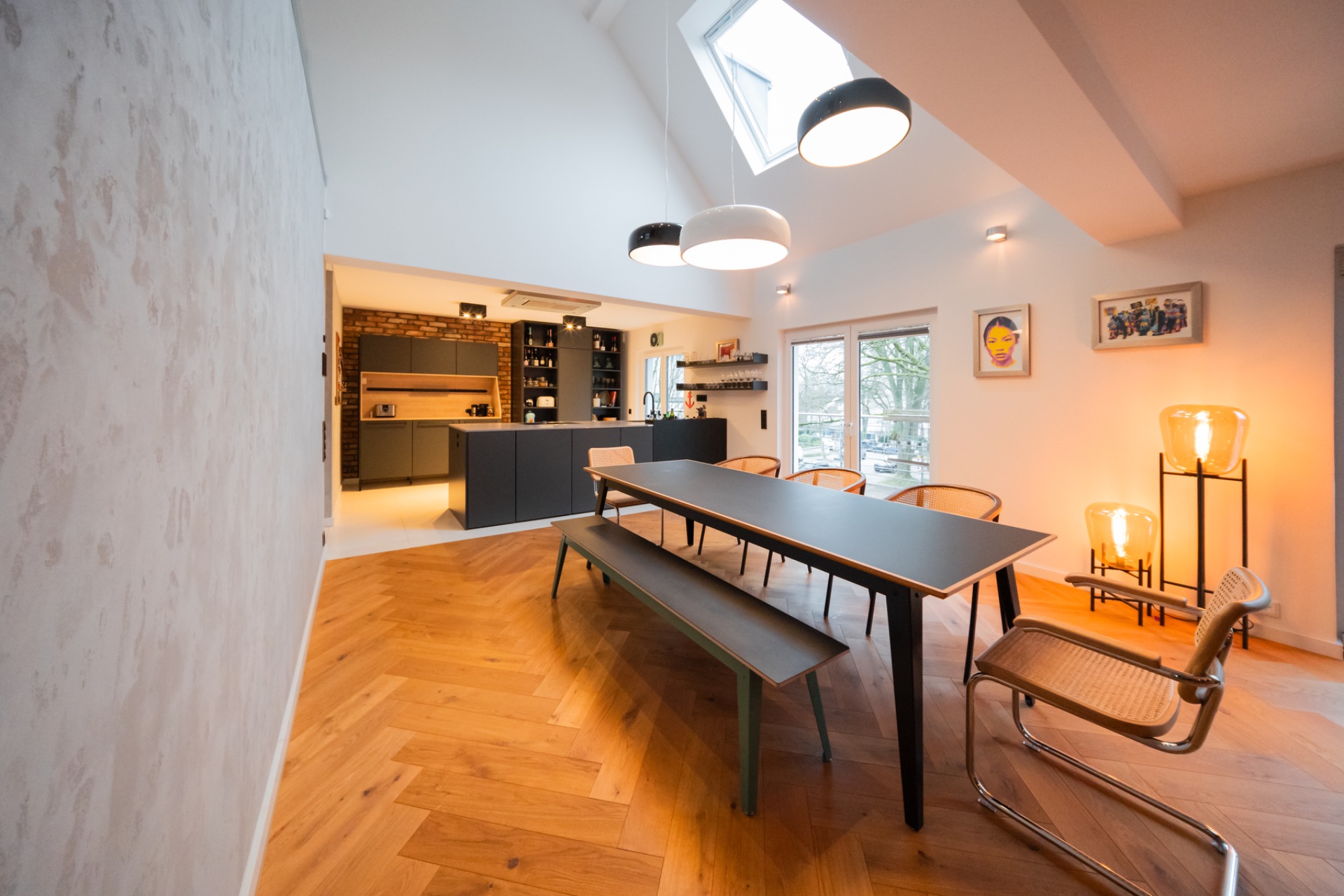 Helles Esszimmer mit langem Tisch, Bank, Stühlen und offener moderner Küche im Hintergrund.