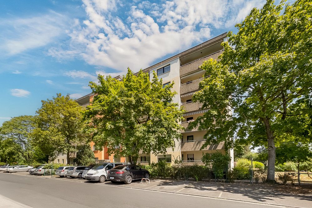 Mehrstöckiges Wohnhaus mit Balkonen, Bäumen davor und parkenden Autos an der Straße.