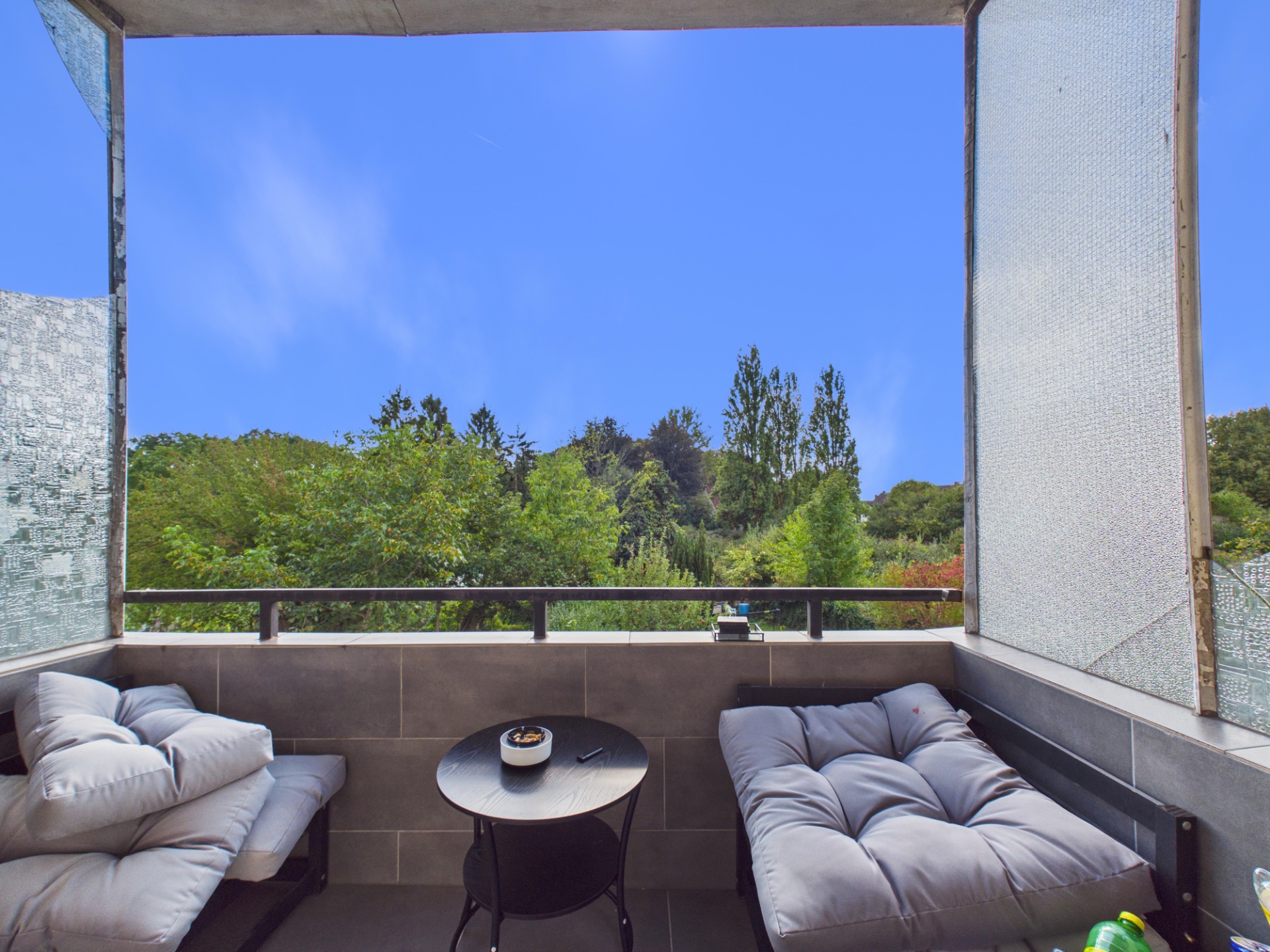 Balkon mit zwei gepolsterten Liegen, kleinem Tisch und Blick auf grüne Bäume.