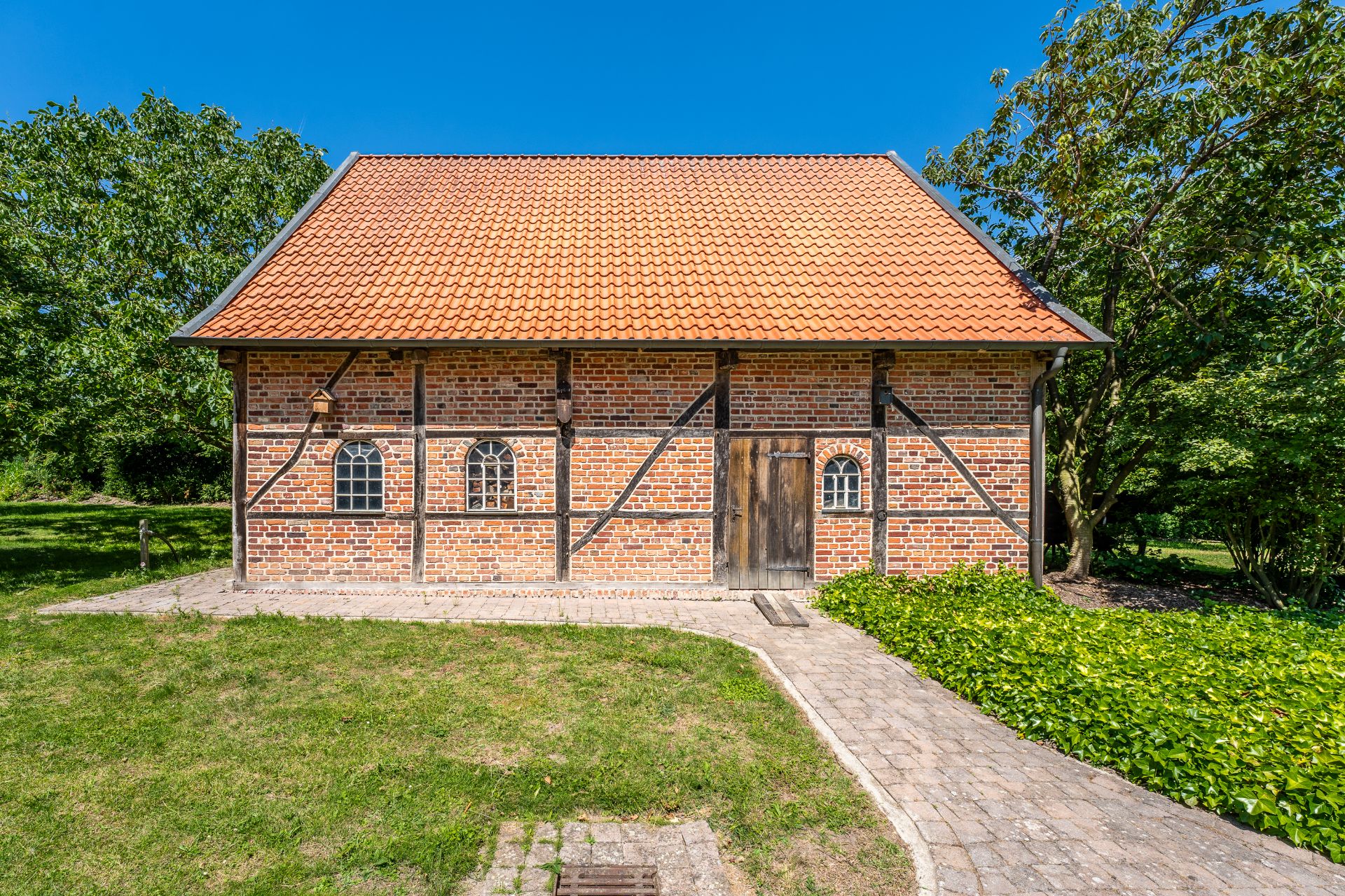 Backstein-Fachwerkhaus mit rotem Ziegeldach, Holztür und drei kleinen Fenstern, umgeben von Garten.