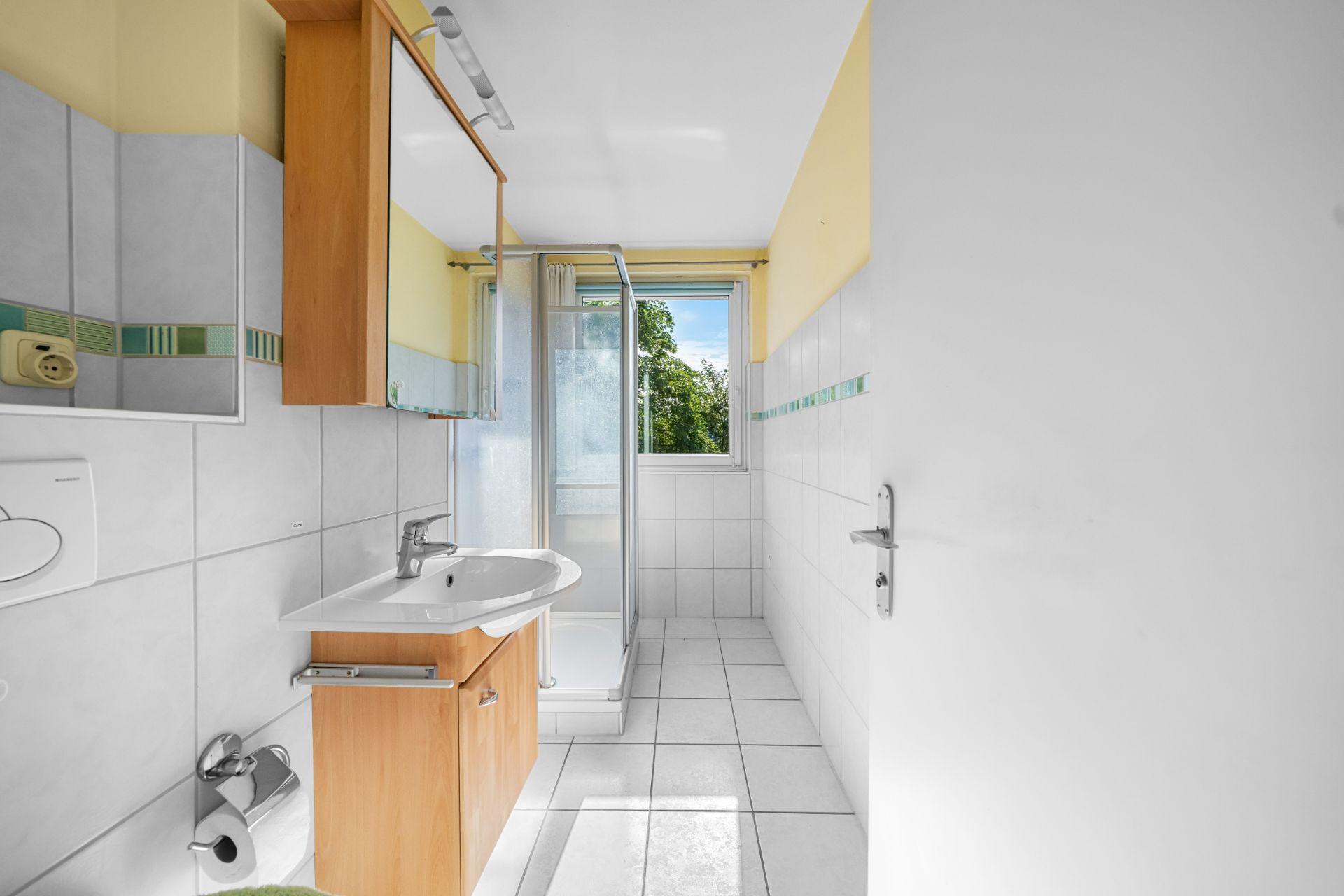 Helles Badezimmer mit Dusche, Waschbecken, Spiegel und Fenster nach außen.