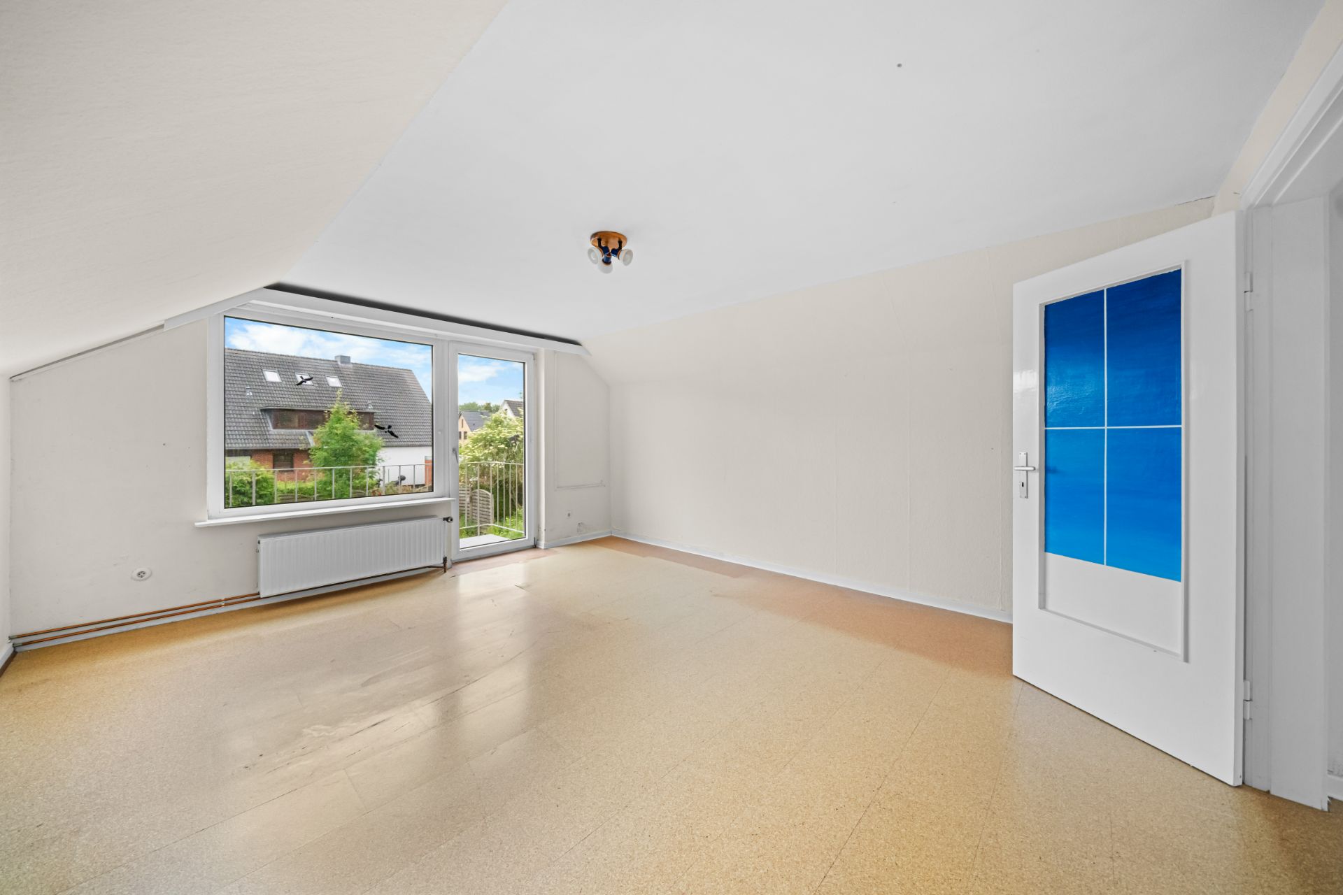 Helles, leeres Zimmer mit großem Fenster, Tür und Blick auf Nachbarhäuser.