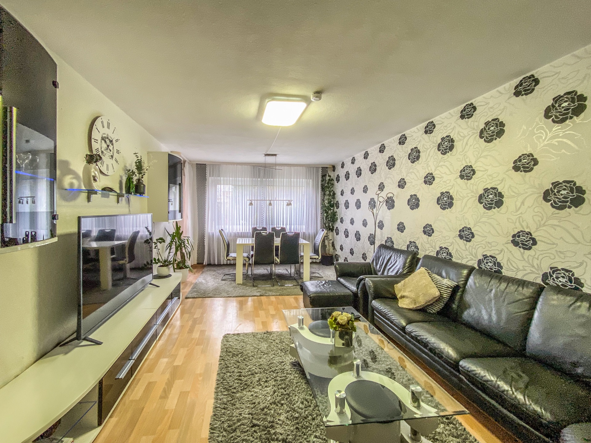 Wohnzimmer mit schwarzem Ledersofa, Glastisch, TV und Essbereich mit sechs Stühlen.