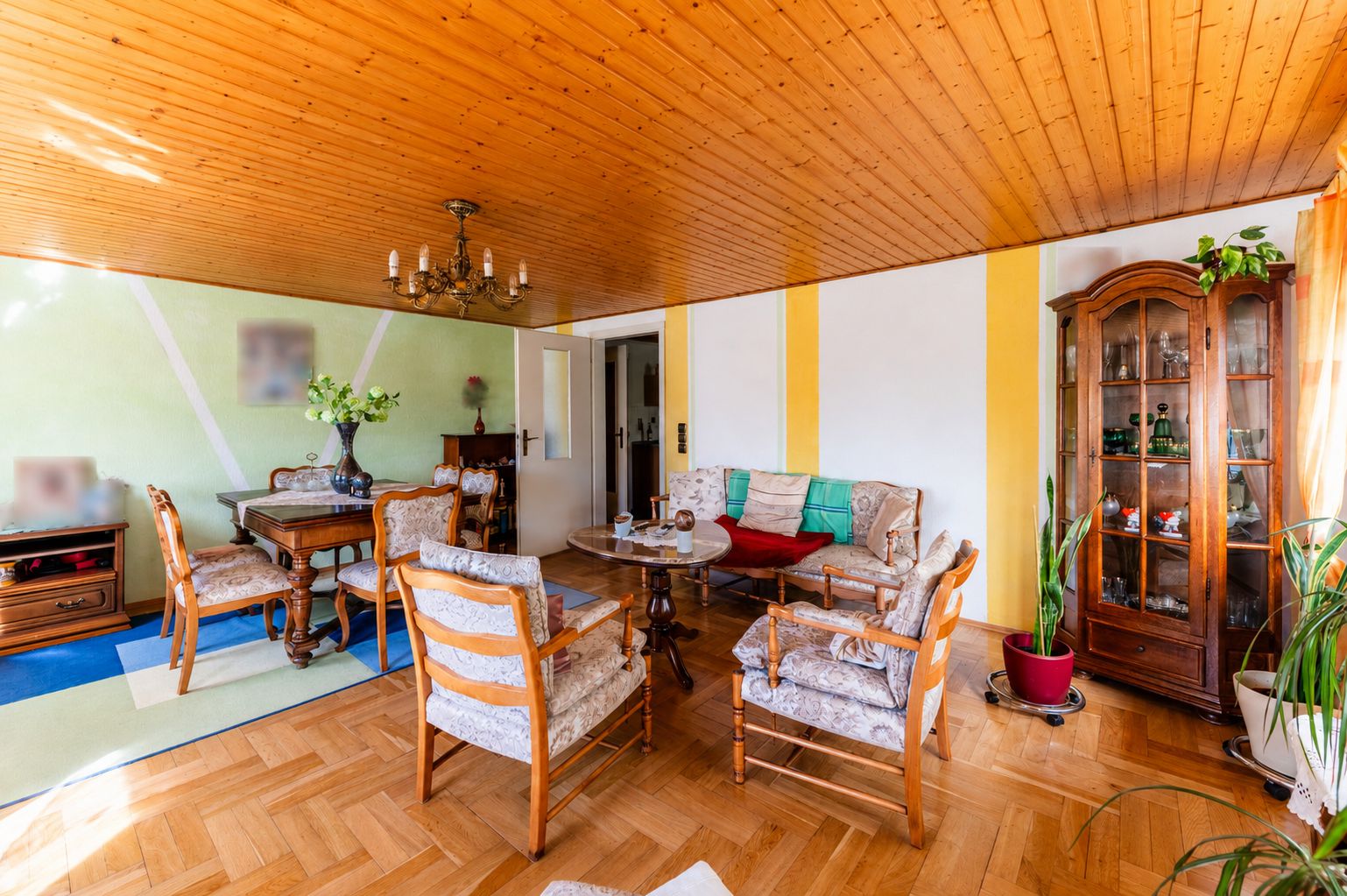 Wohnzimmer mit Holzboden, Sofa, Sesseln, Esstisch, Vitrine und Zimmerpflanzen.