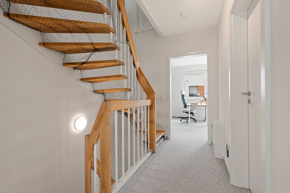 Heller Flur mit Holztreppe und Blick in ein Büro mit Schreibtisch und Stuhl.