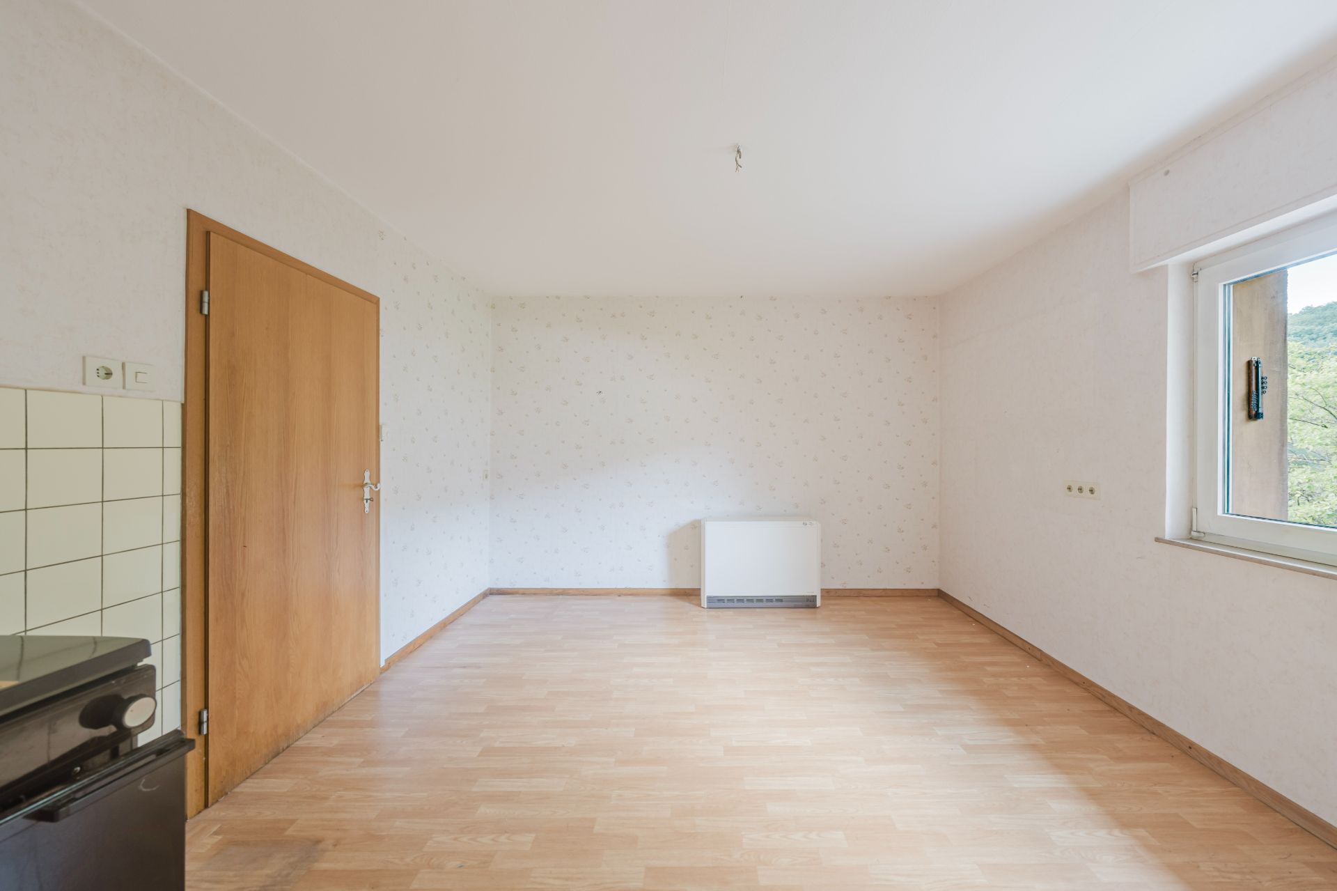 Leeres Zimmer mit Holzboden, Fenster rechts, Holztür links und einem Heizkörper an der Rückwand.