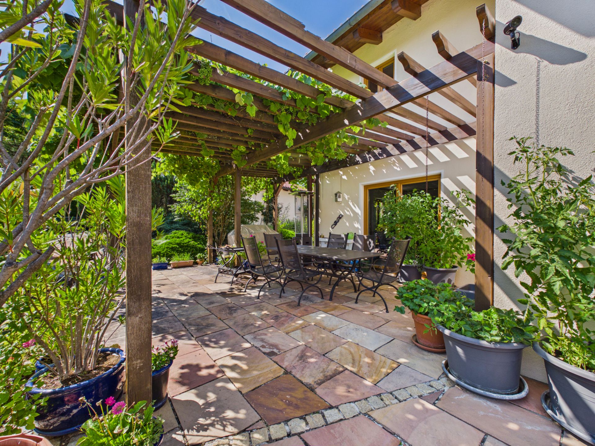Überdachte Terrasse mit Esstisch, Stühlen und vielen Pflanzen in Töpfen neben einem Haus.