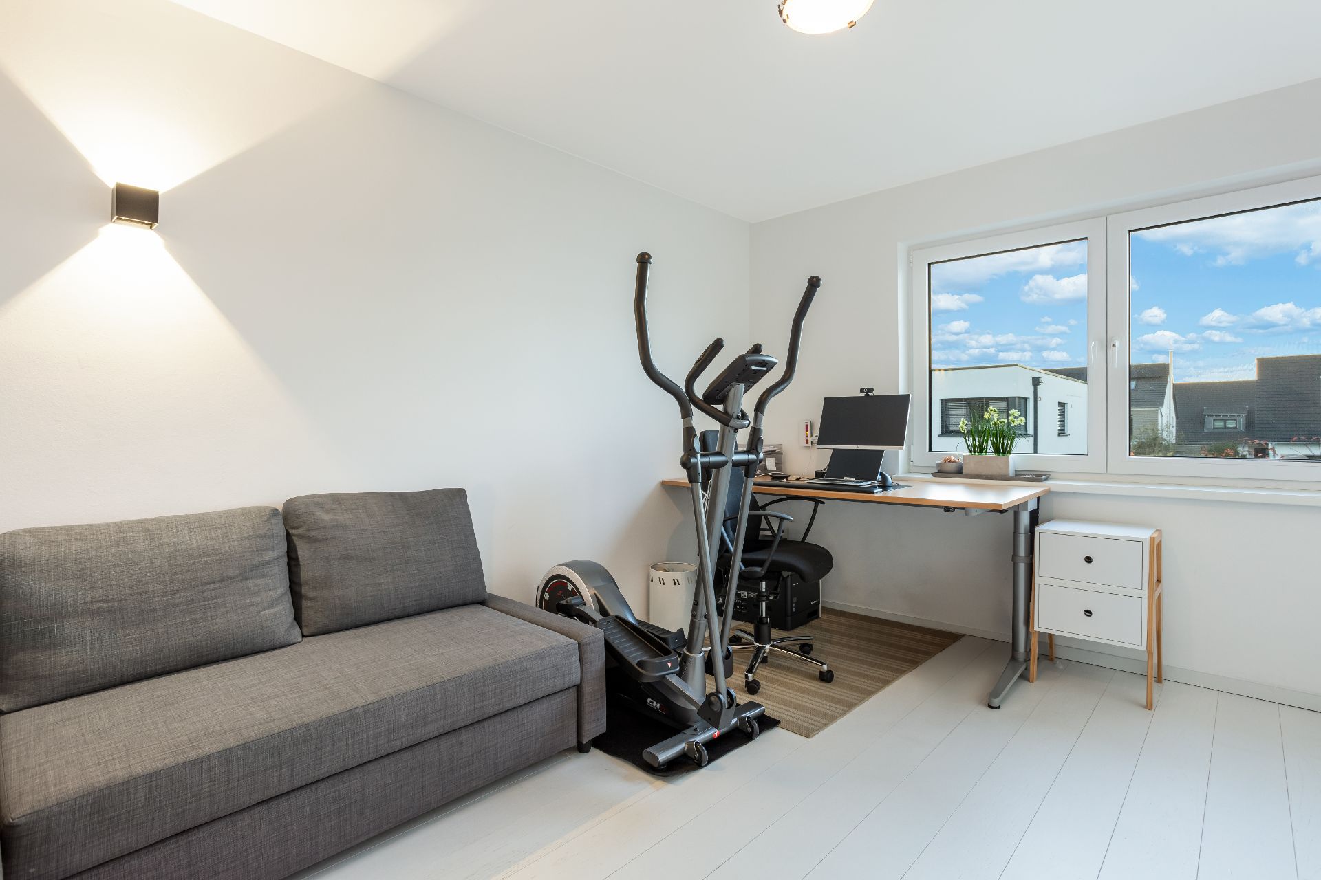 Helles Zimmer mit grauem Sofa, Schreibtisch, Computer und Crosstrainer, großes Fenster mit Ausblick.