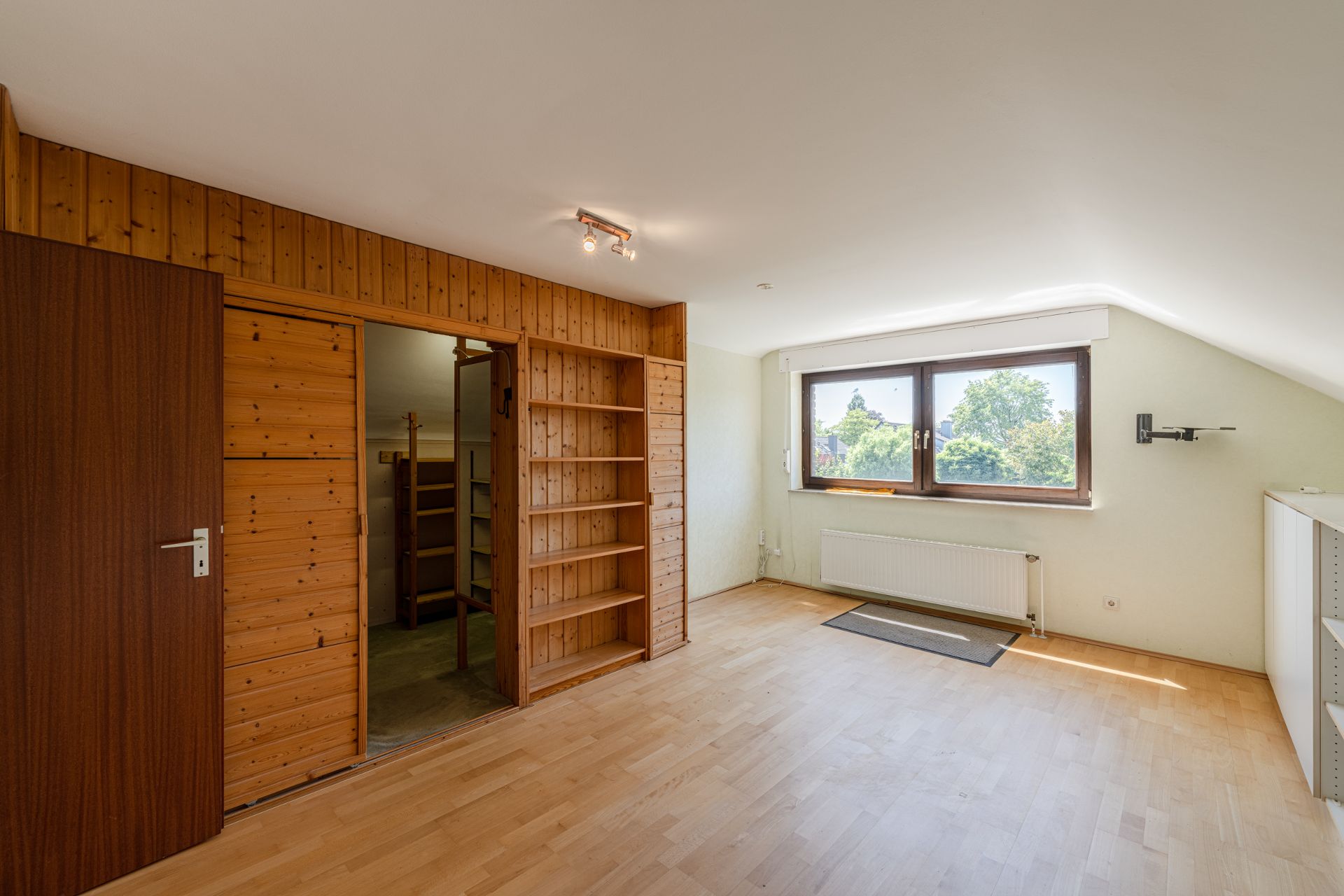 Helles Zimmer mit Holzregalen, Holztüren, großen Fenstern und Heizkörper unter dem Fenster.