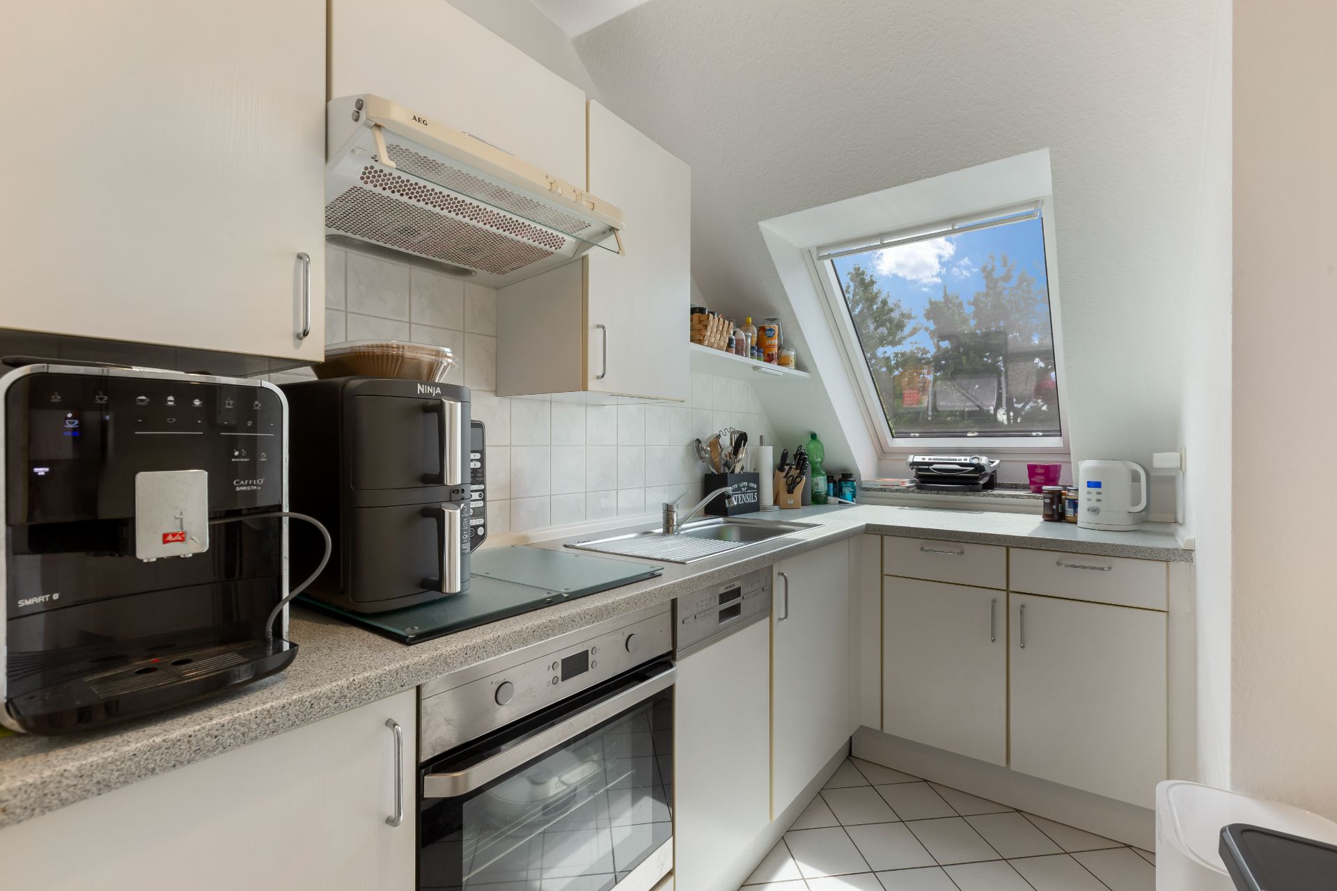Moderne Einbauküche mit weißen Schränken, Kaffeemaschine und großem Fenster unter einer Schräge.
