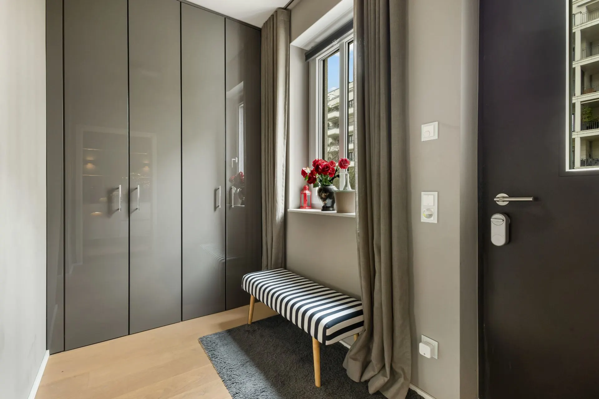Flur mit grauem Einbauschrank, gestreifter Bank und Fenster mit Blumen auf der Fensterbank.
